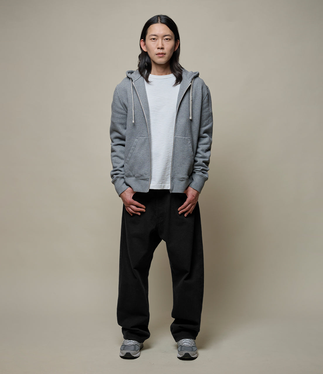 Person wearing a gray hoodie, white t-shirt, black pants, and gray shoes on a beige background