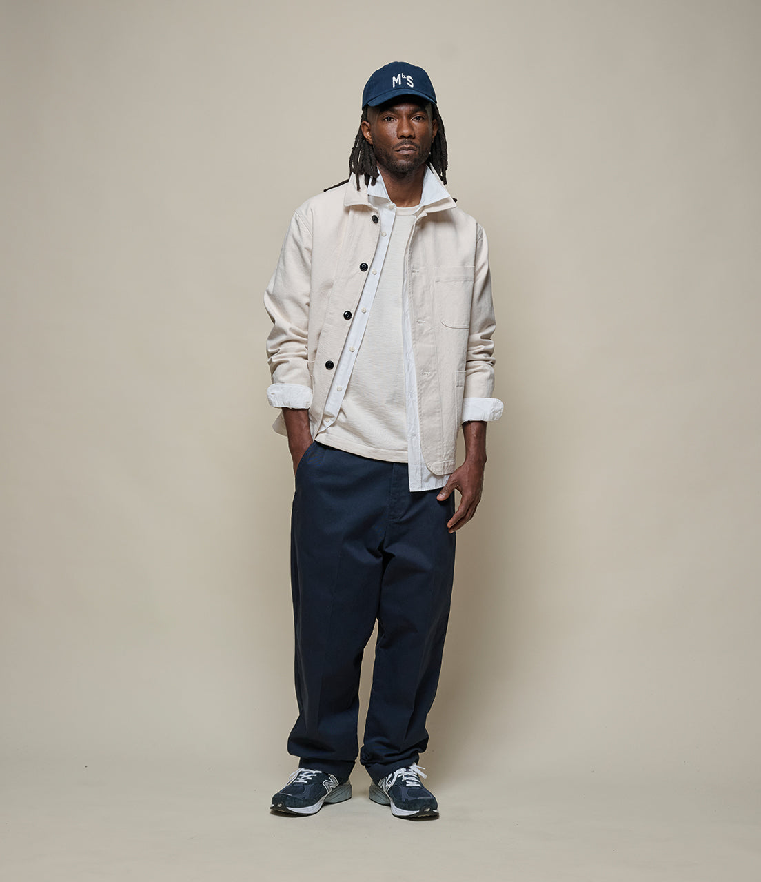 Man wearing a beige jacket, white shirt, navy pants, and blue cap on a beige background
