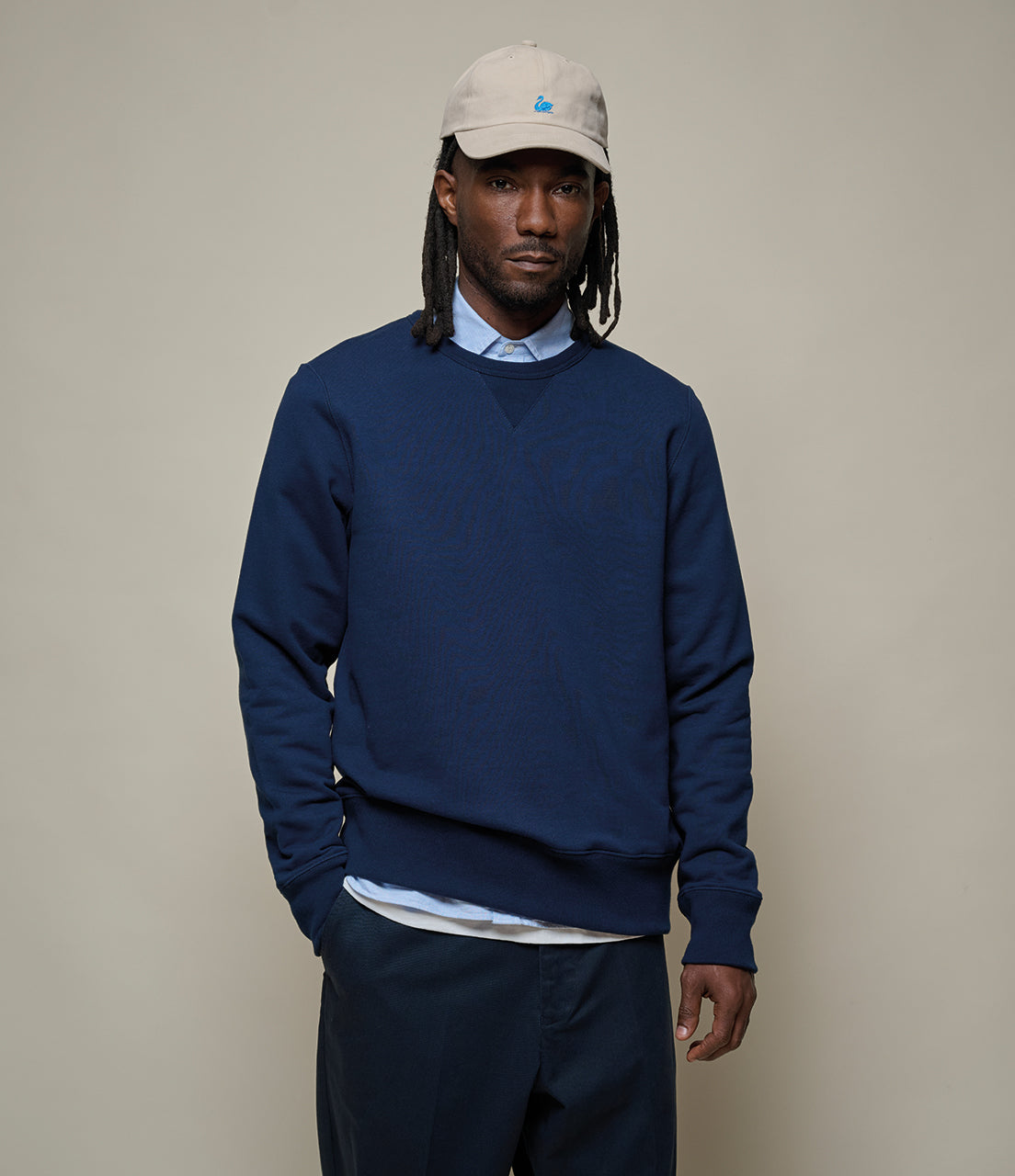 Man wearing a navy blue sweater, beige cap, and dark pants against a plain background
