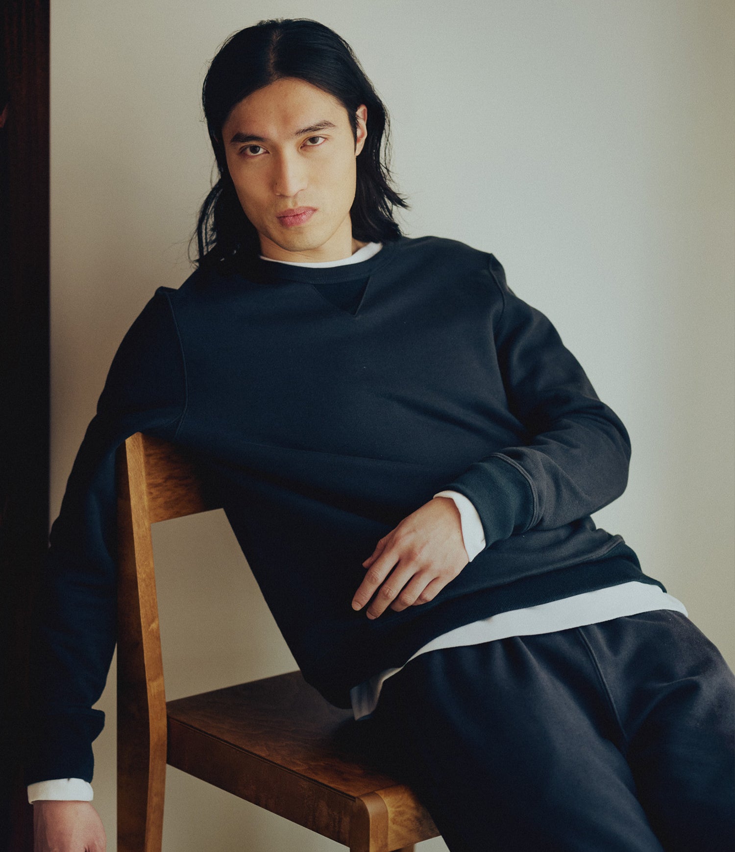 A man with long dark hair sits on a wooden chair wearing a dark grey crewneck sweatshirt over a white shirt, paired with dark navy sweatpants against a neutral indoor background.
