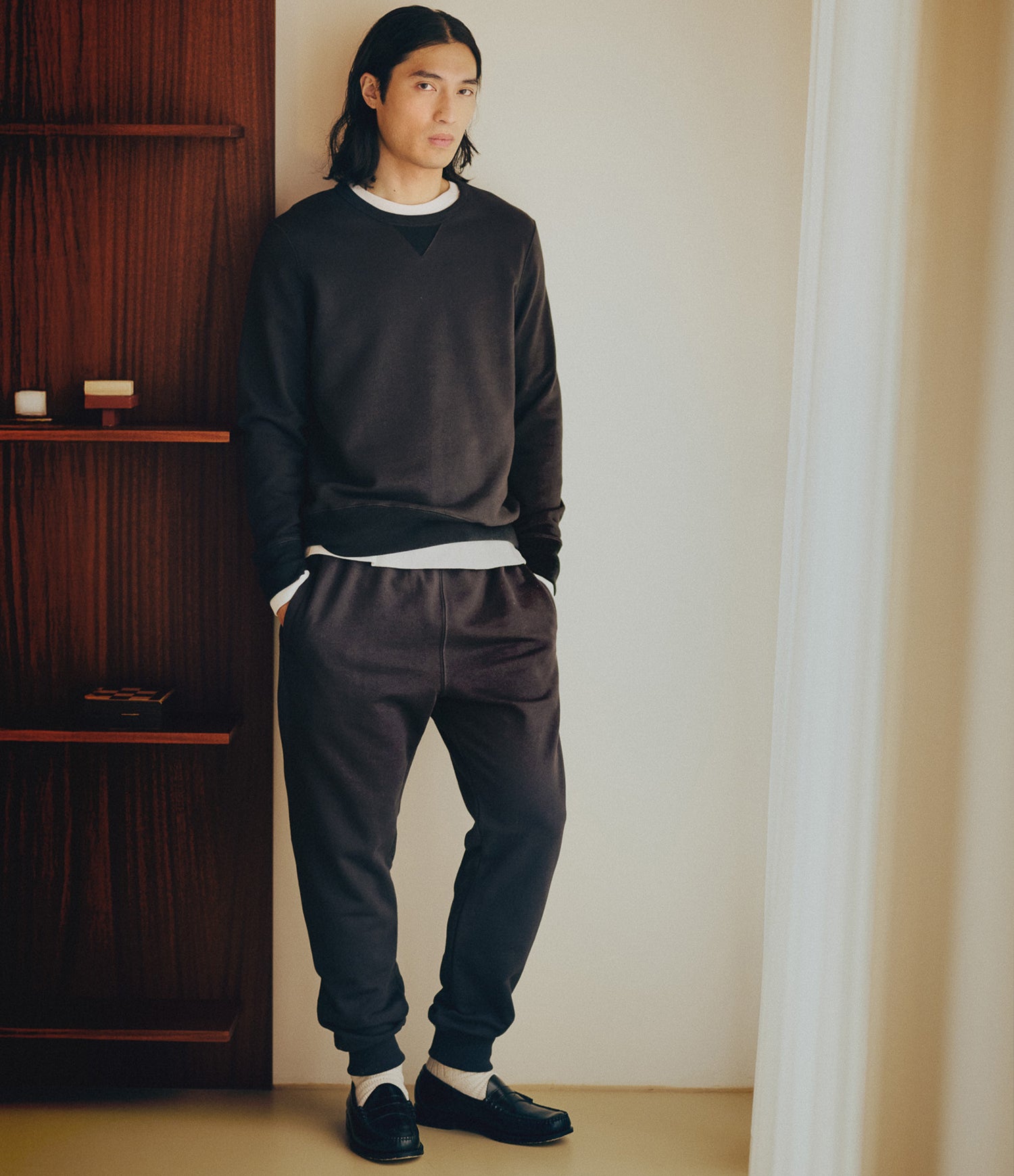A man wearing a dark charcoal gray v-neck sweatshirt, matching jogger sweatpants, white undershirt, white socks, and black loafers, leaning against a beige wall next to a dark wooden shelf.