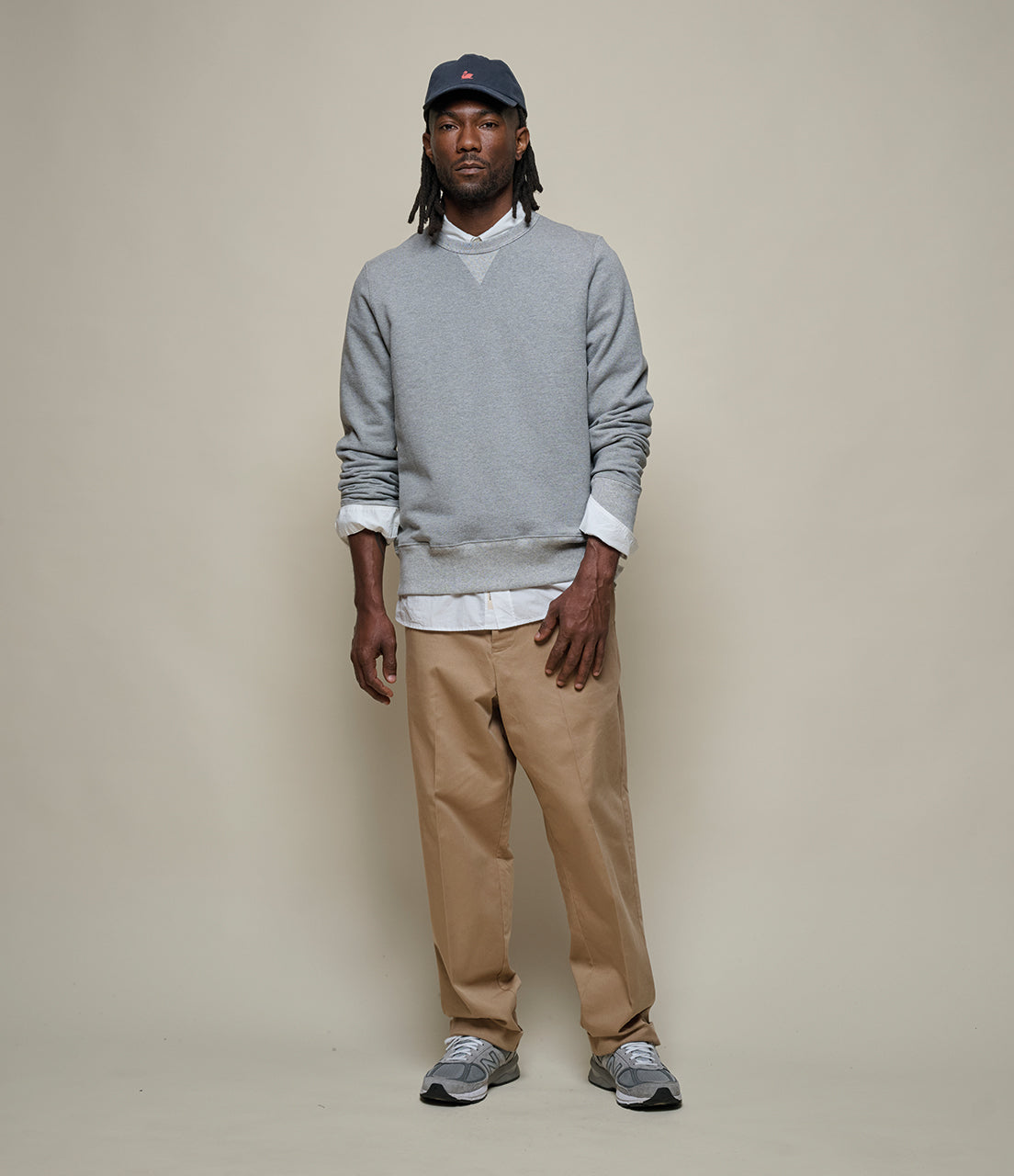 Man wearing a gray sweater, beige pants, and a cap against a plain background