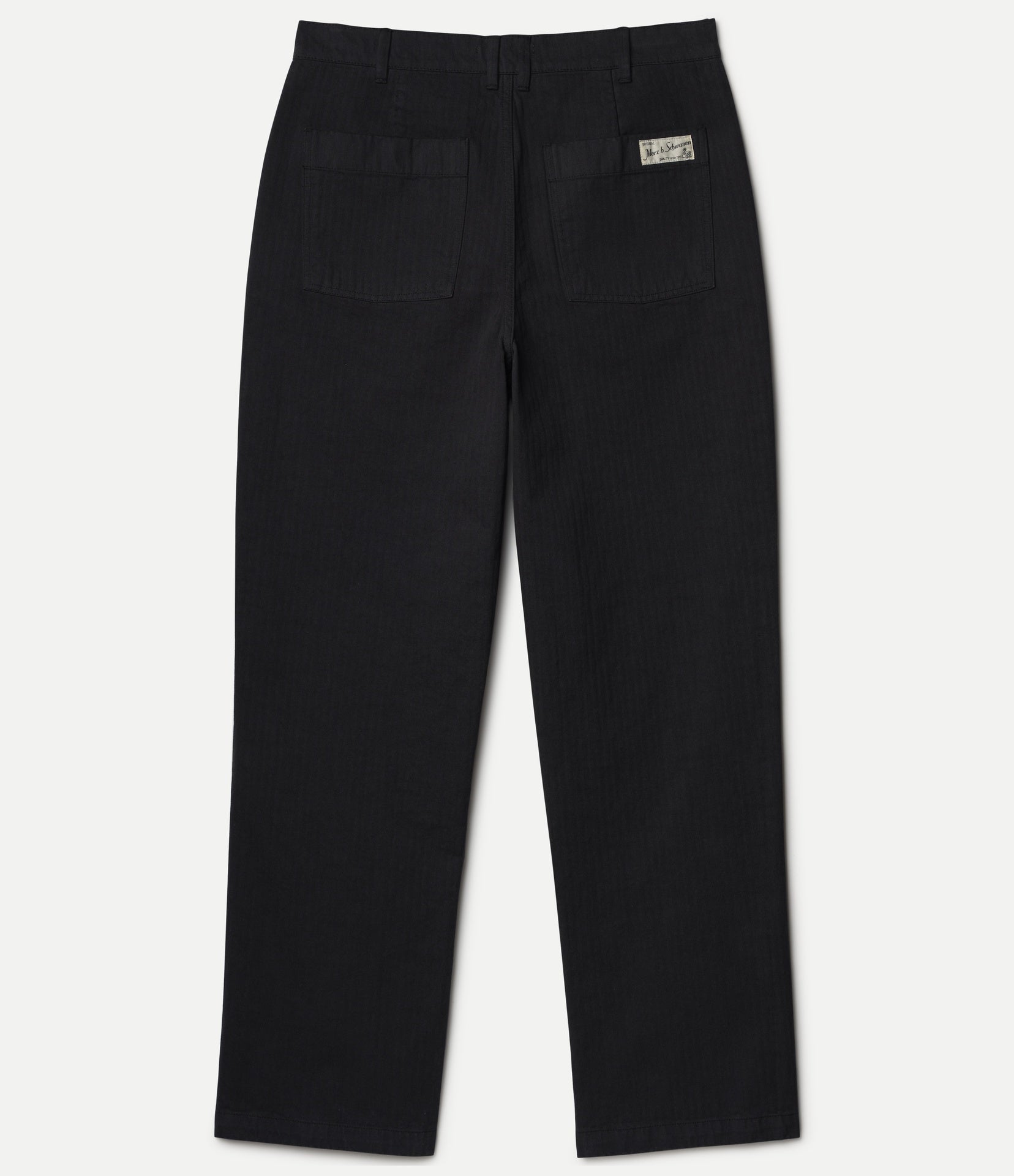 Black straight-leg pants shown from the back, featuring two back pockets and a small rectangular Merz b. Schwanen brand patch on the upper right side, set against a light off-white background.