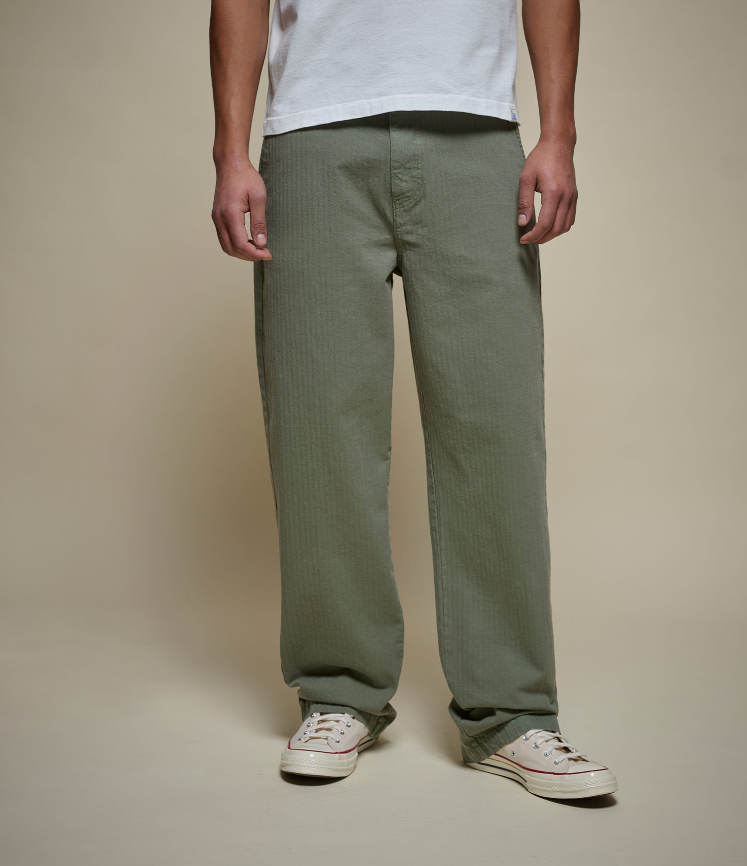 A person wearing straight-leg olive green herringbone pants, styled with a white t-shirt and white low-top sneakers, standing against a solid beige studio background.