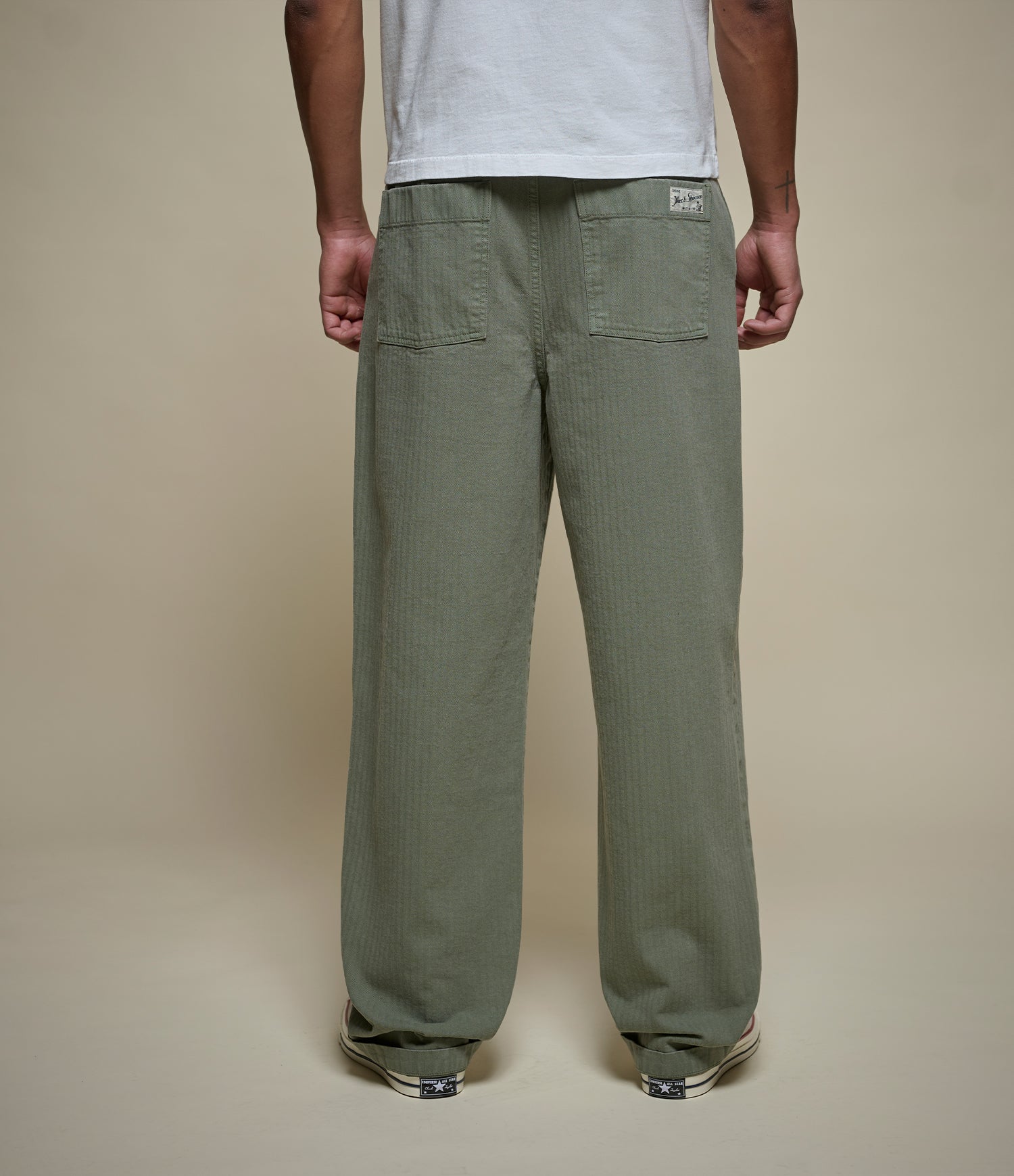 A person shown from the back wearing loose, straight-leg olive green herringbone pants and a white t-shirt against a plain beige studio background.