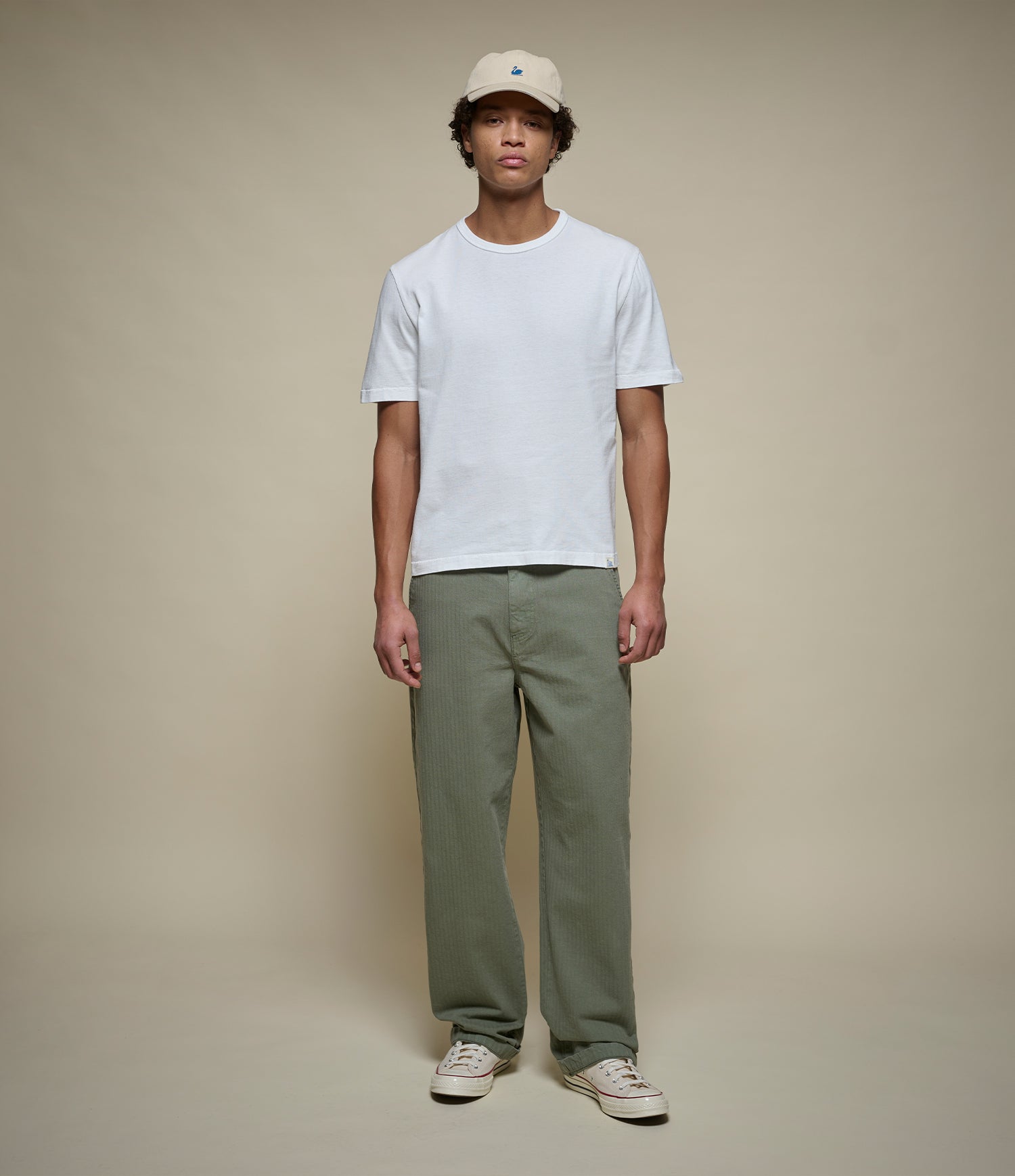 A man modeling a white short-sleeve crewneck t-shirt, olive green straight-leg pants, and a beige baseball cap against a solid beige studio background.