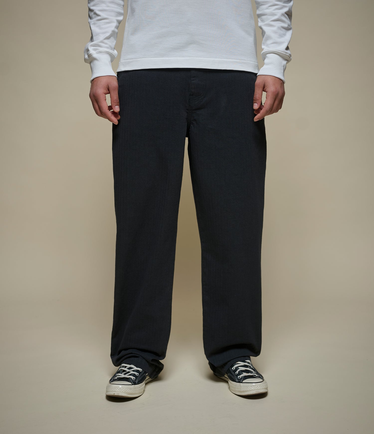 A person wearing loose black straoght-leg pants with herringbone pattern, a white long-sleeve shirt, and black low-top sneakers, standing against a plain beige studio background.