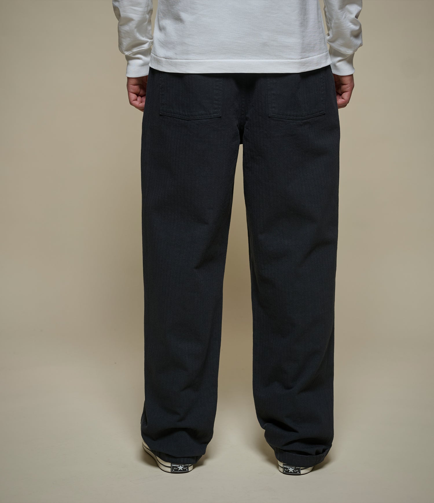 A person shown from the back wearing loose, straoght-leg dark herringbone pants and a white long-sleeve top against a plain beige studio background.