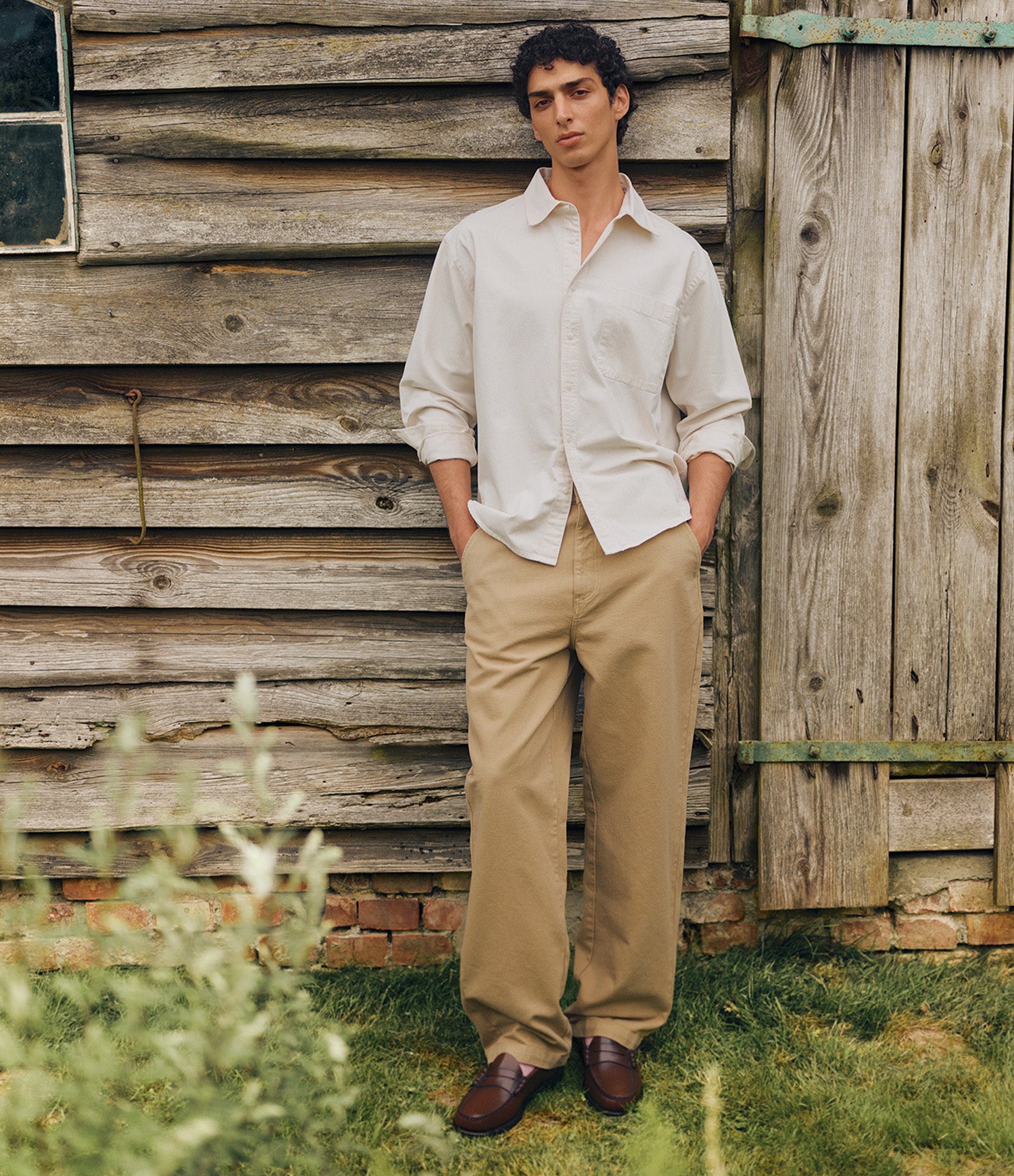 A man stands outdoors in front of a rustic wooden cabin wall, wearing a light cream button-down shirt, tan trousers, and brown leather shoes.