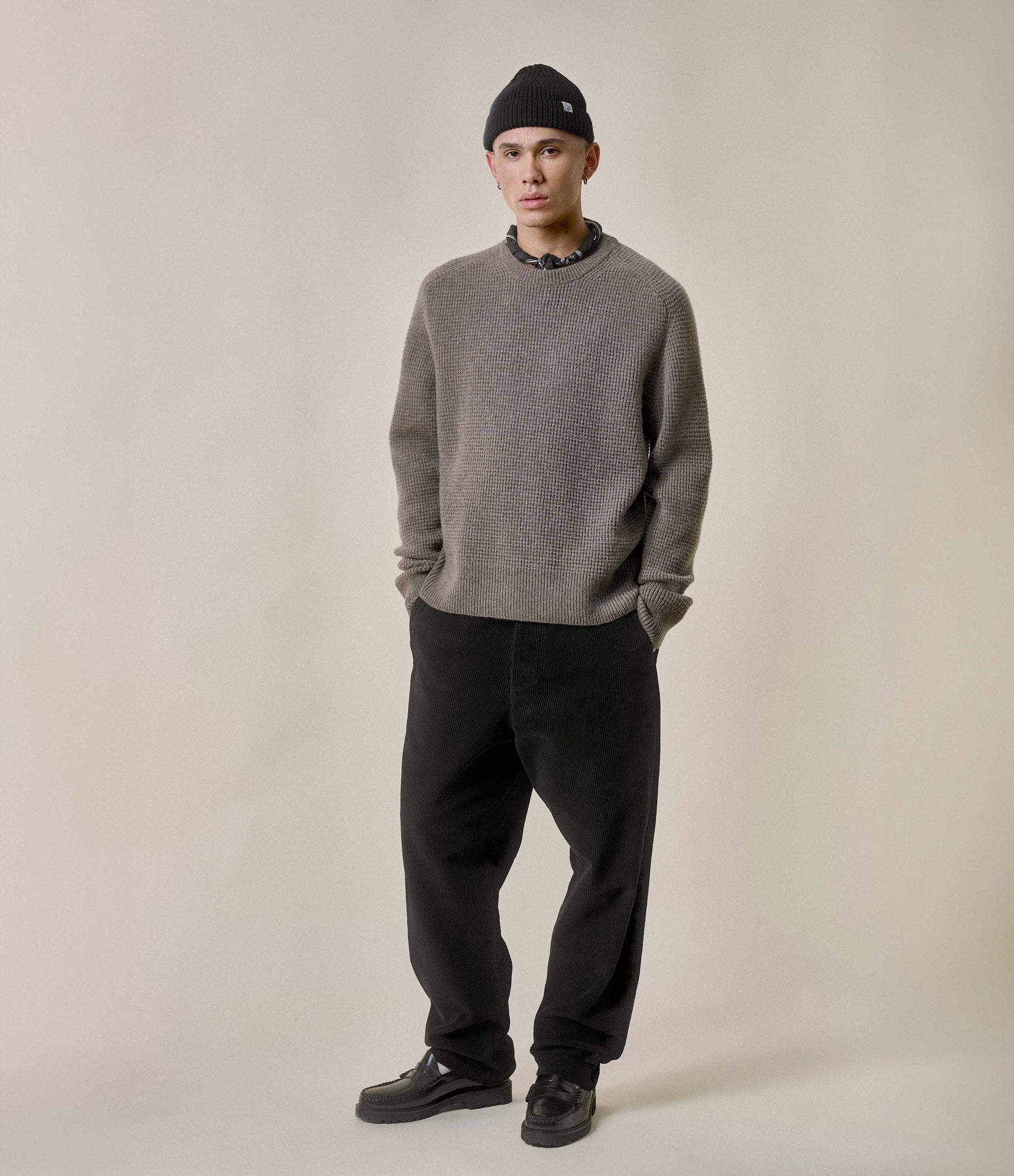 Man wearing a gray sweater, black pants, and a black beanie against a plain background