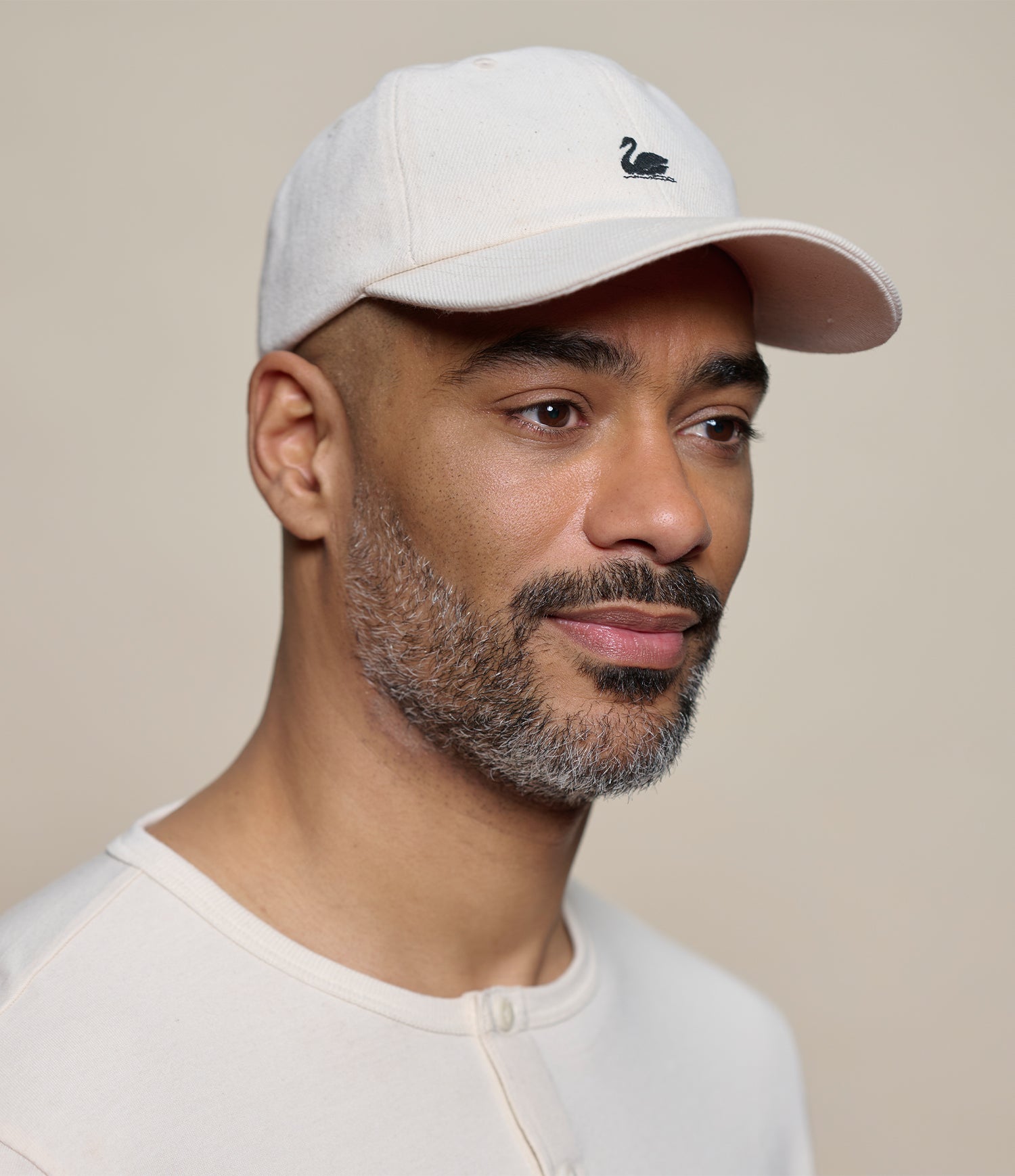 A close-up portrait of a man wearing a light beige baseball cap with a small black swan logo on the front, paired with a matching light beige henley shirt against a solid beige background.