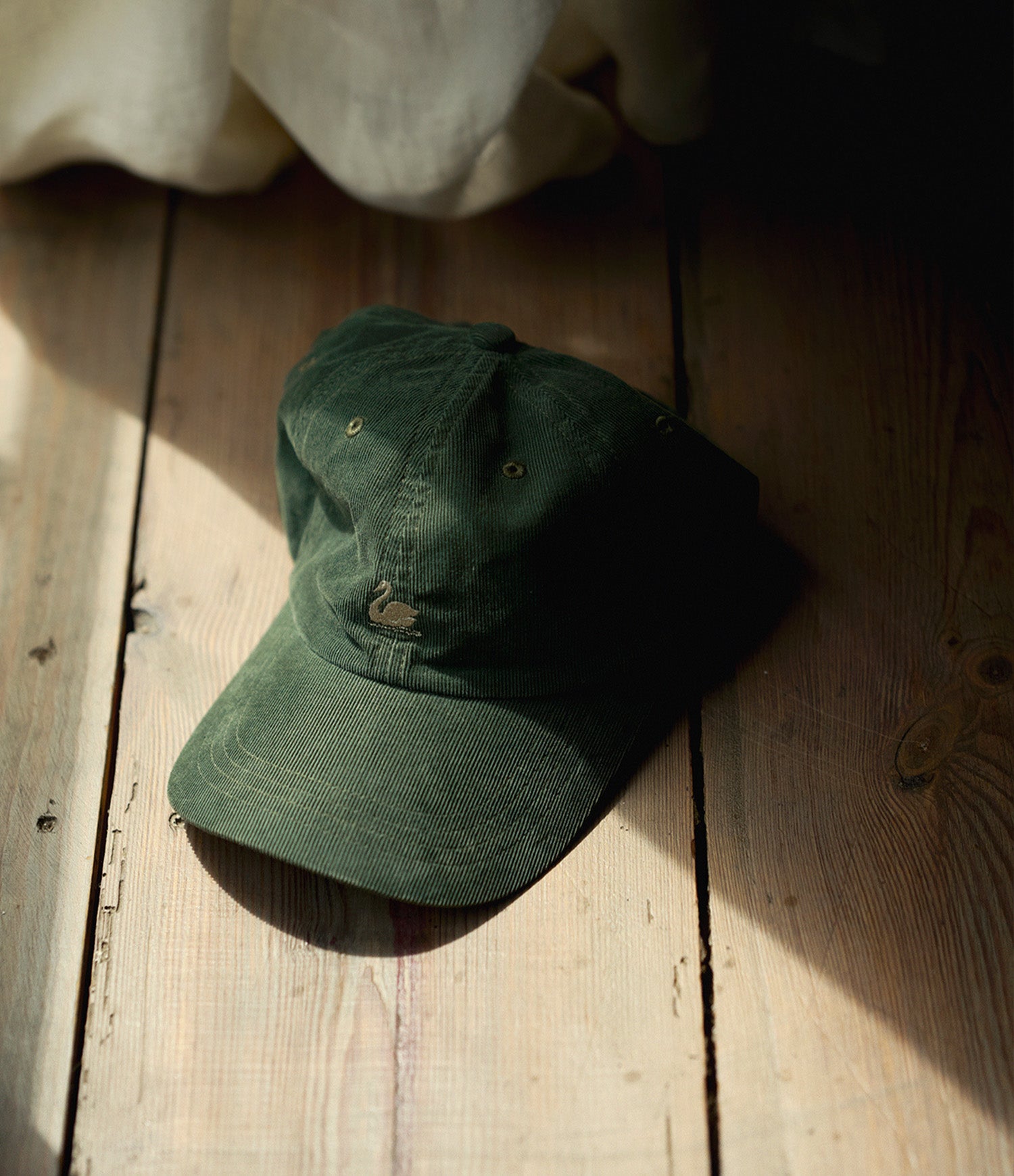 A dark green cotton baseball cap with a small embroidered swan logo, resting on a rustic wooden plank floor with dramatic sunlight and shadows.