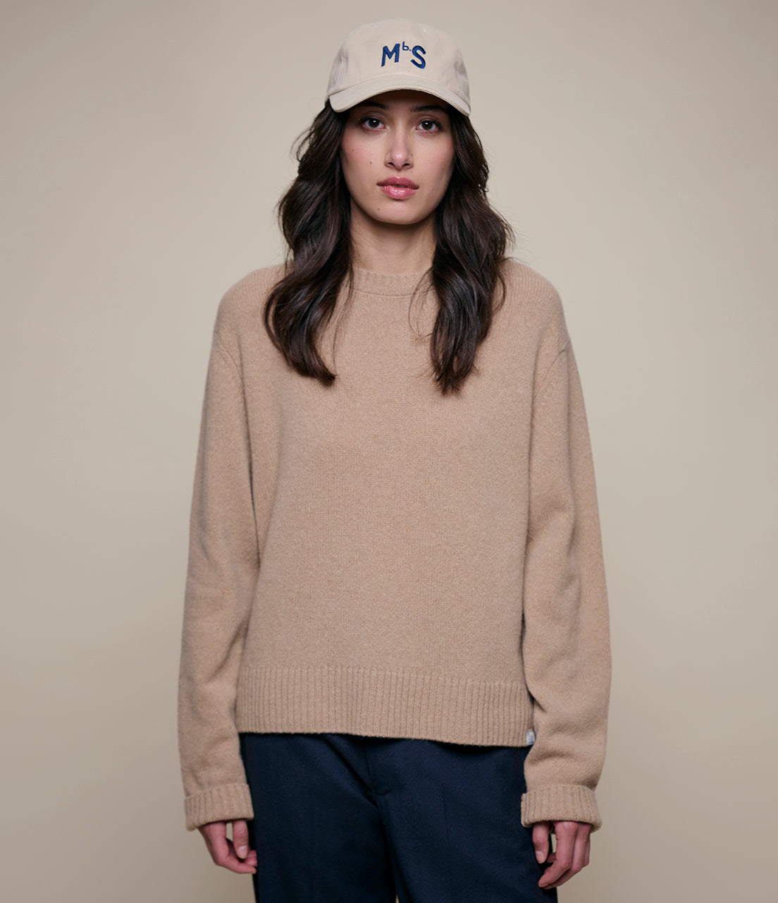 Woman wearing a beige cashmere sweater and a cap with 'MbS' logo on a beige background