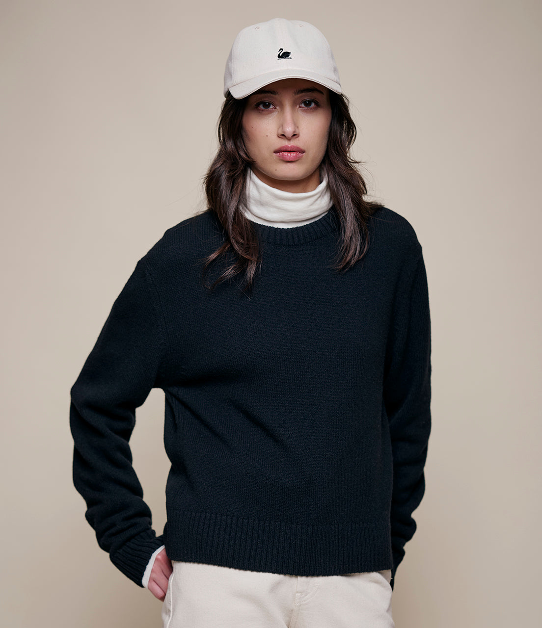 Person wearing a black cashmere sweater, white turtleneck, and white cap on a beige background