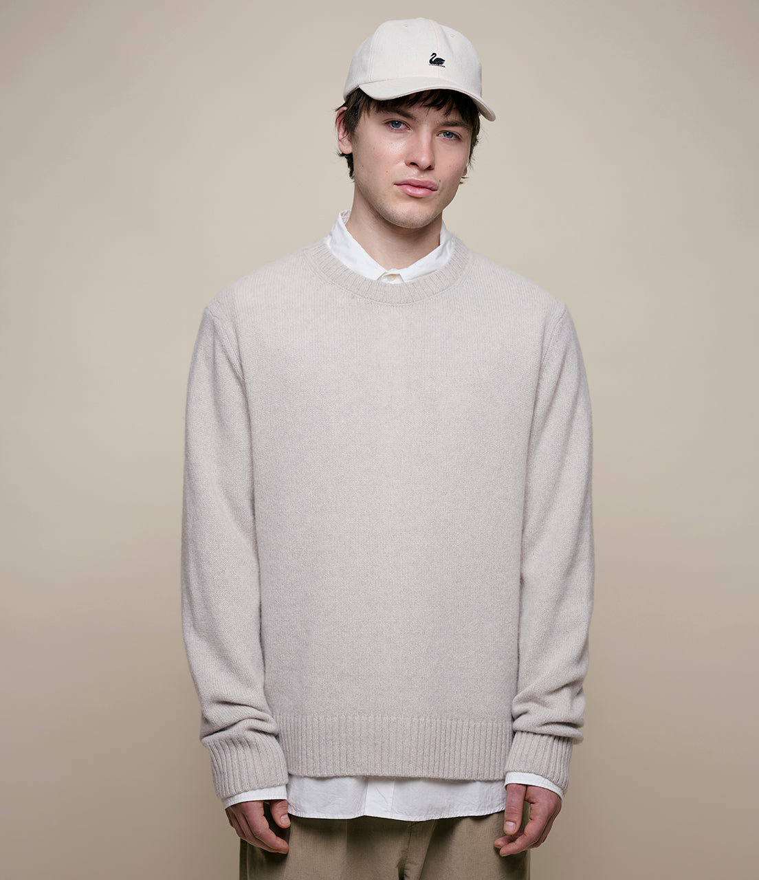 Person wearing a light gray cashmere sweater over a white shirt with a beige cap on a plain background