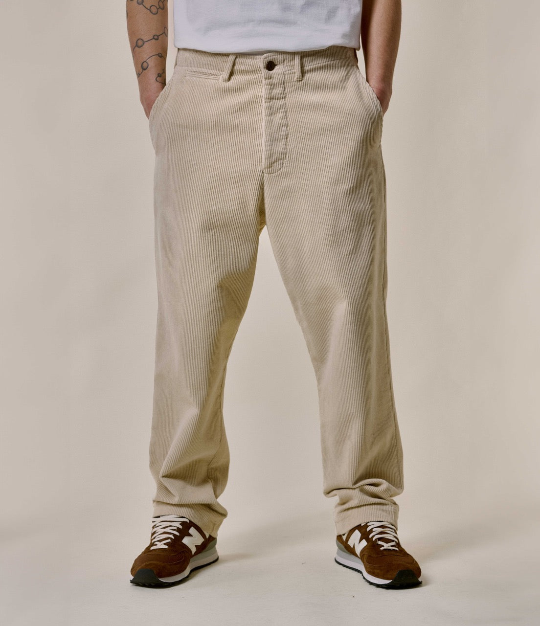 A person standing against a plain background, wearing beige corduroy chino pants with a relaxed fit and pleats, paired with a white T-shirt and brown sneakers.