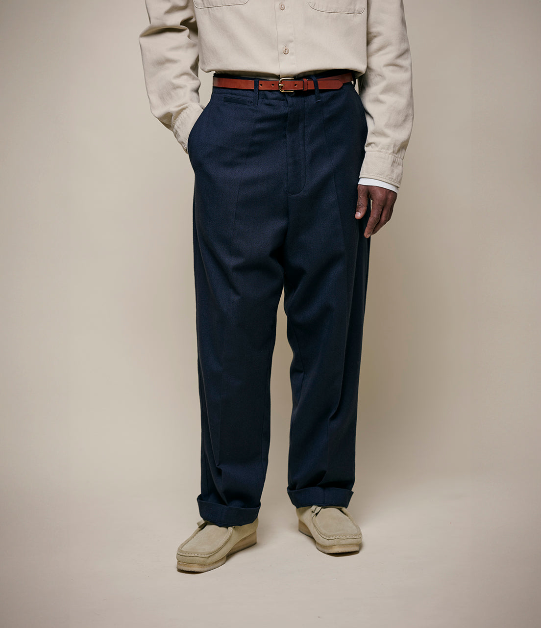 Person wearing navy wool pants and a beige shirt with a brown belt on a plain background