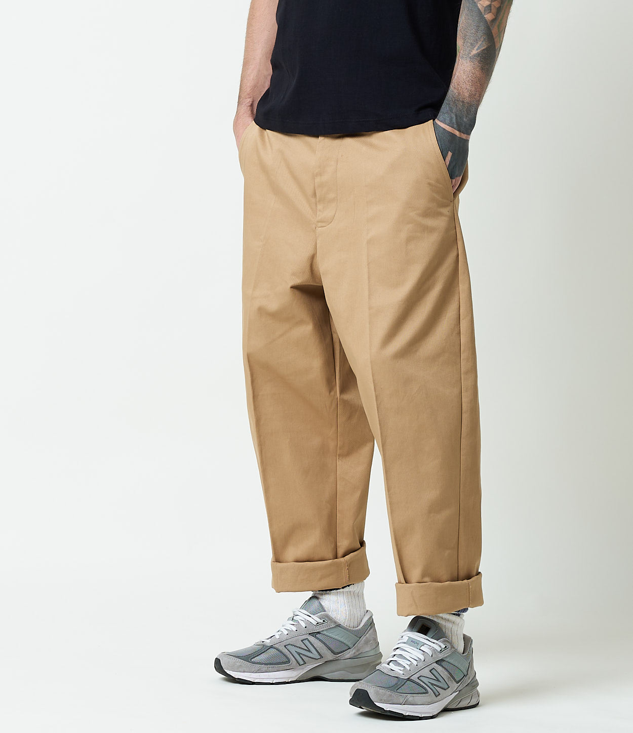 A person standing against a white background, wearing beige chino pants with a rolled-up hem and a pair of gray and black sneakers.