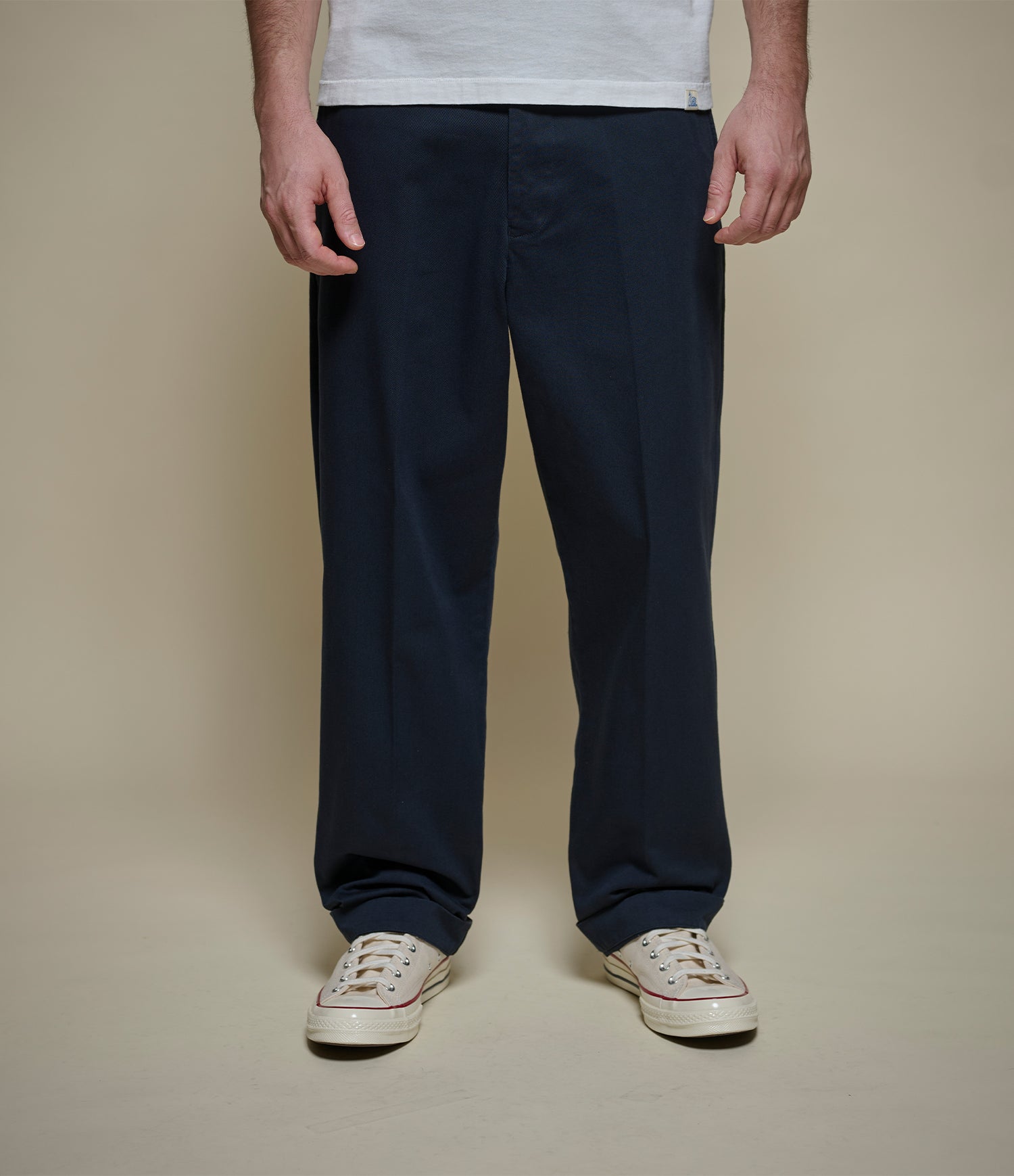 A person wearing dark navy blue straight-leg chino, a white t-shirt, and white canvas sneakers, standing against a solid beige studio background.