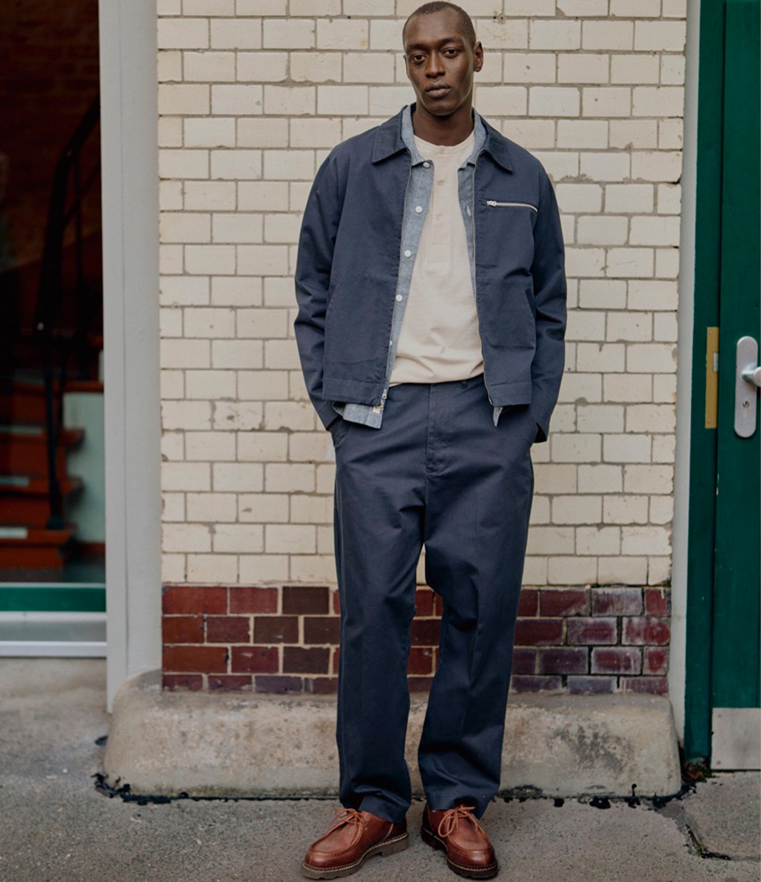 A man stands outdoors against a cream brick wall wearing a navy blue work jacket and matching navy pants over a cream t-shirt, with brown leather shoes.