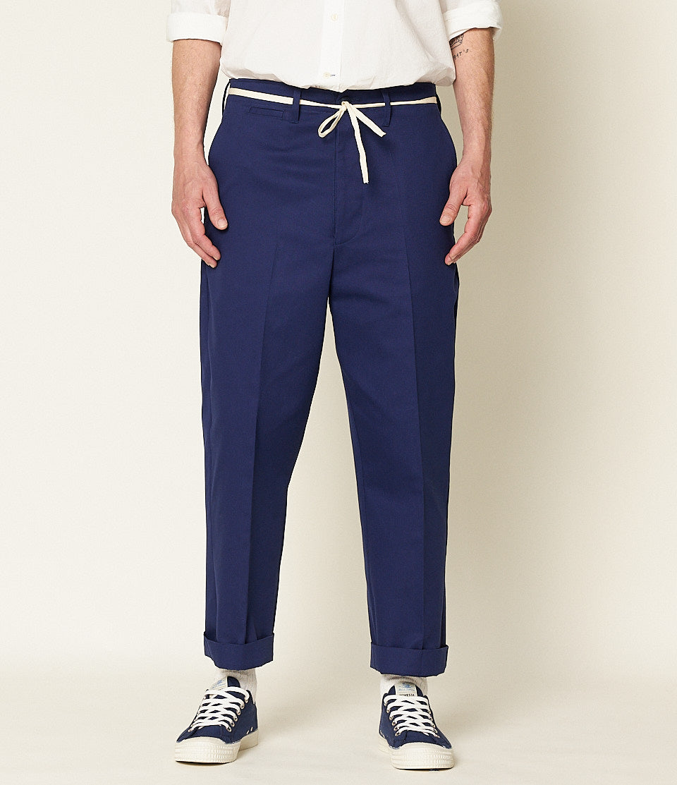 A person standing against a beige background, wearing navy blue chino pants with a white belt and white sneakers.