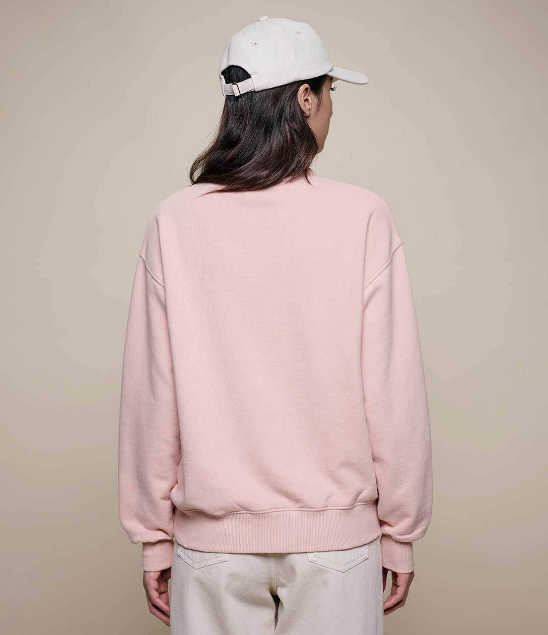 Person wearing a light pink sweatshirt and beige pants with a white cap on a beige background