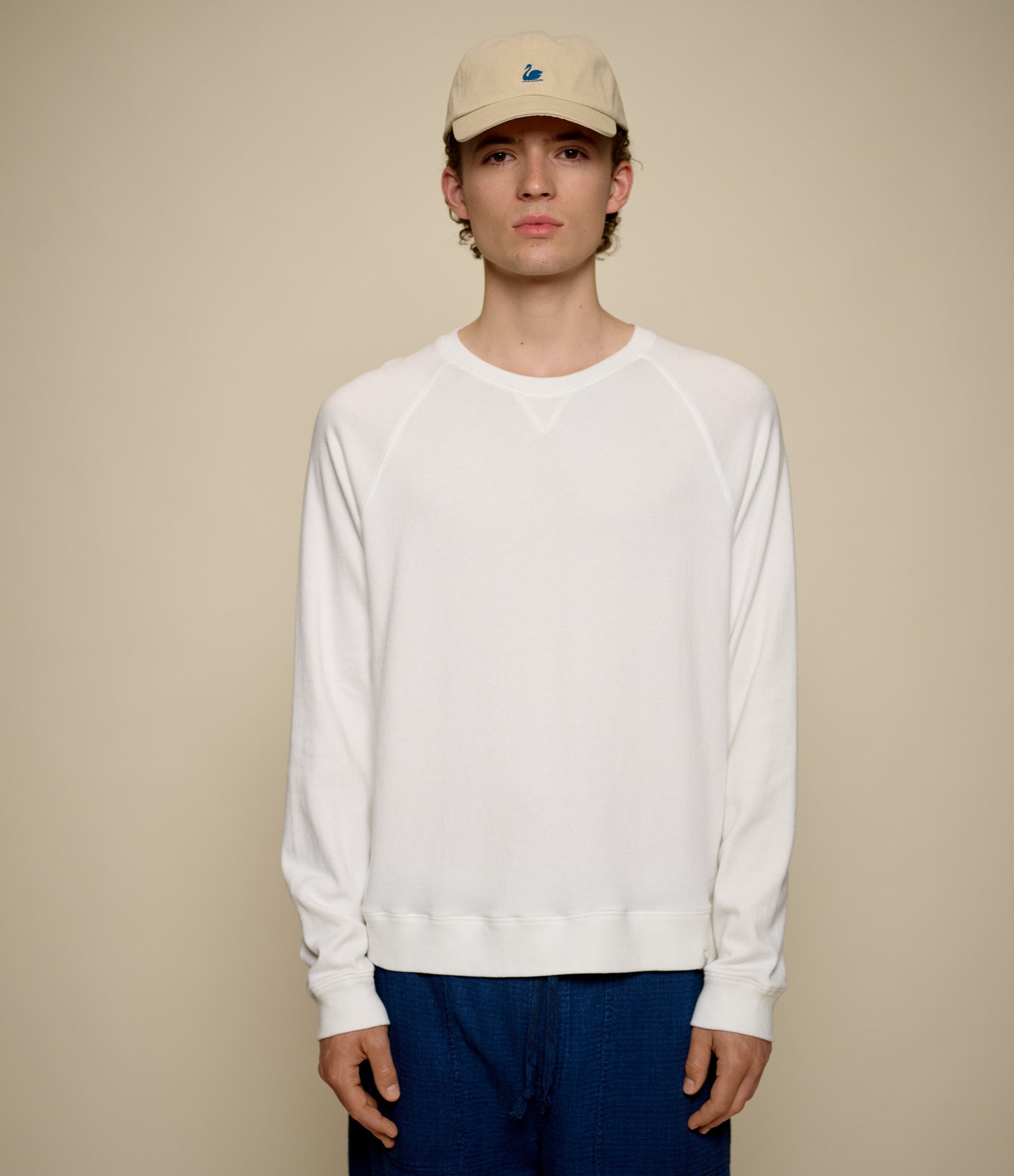 Person wearing a white lightweight sweatshirt and blue pants with a beige cap on a beige background
