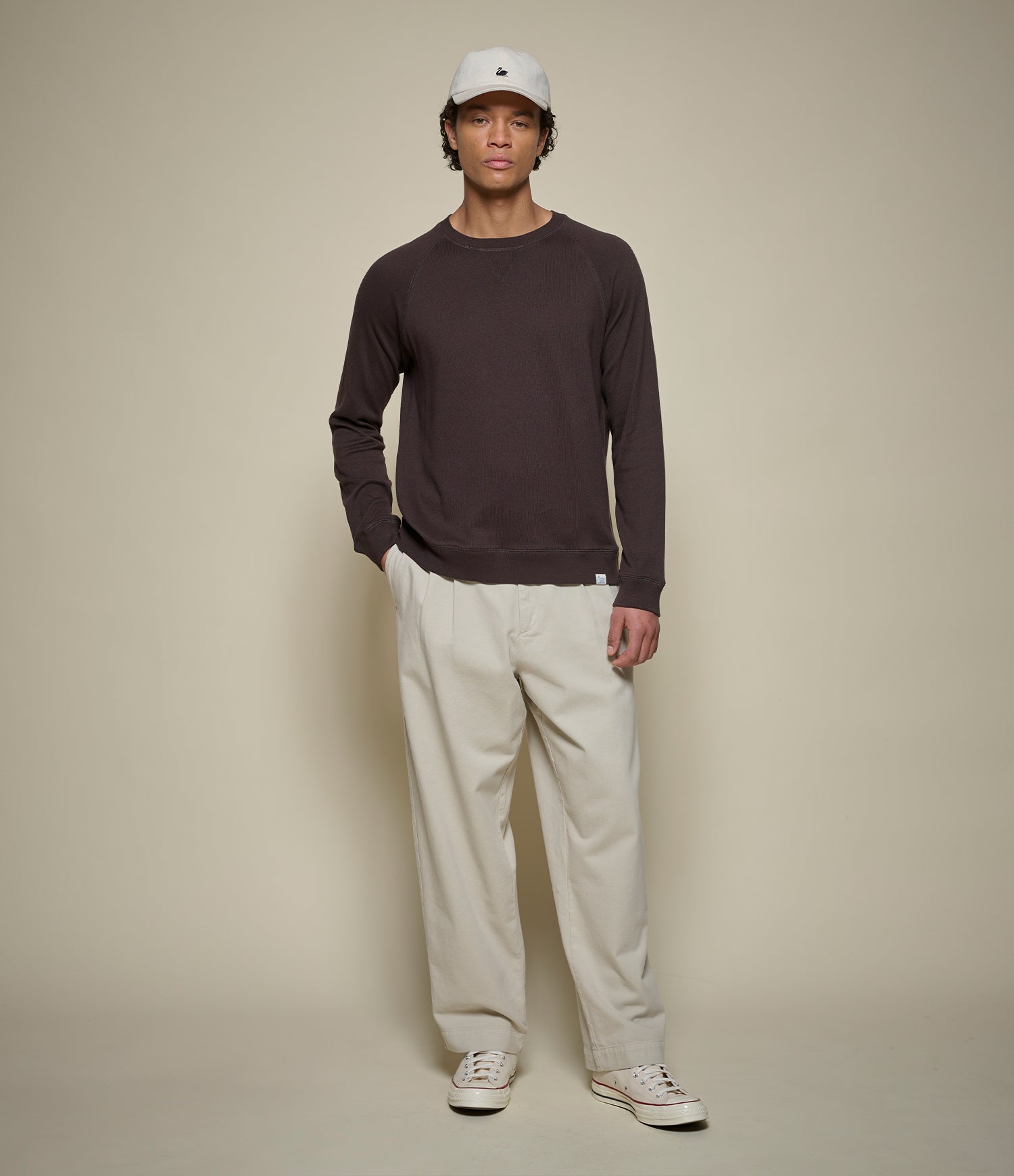 Man wearing a brown lightweight sweatshirt, beige pants, and a white cap against a beige background