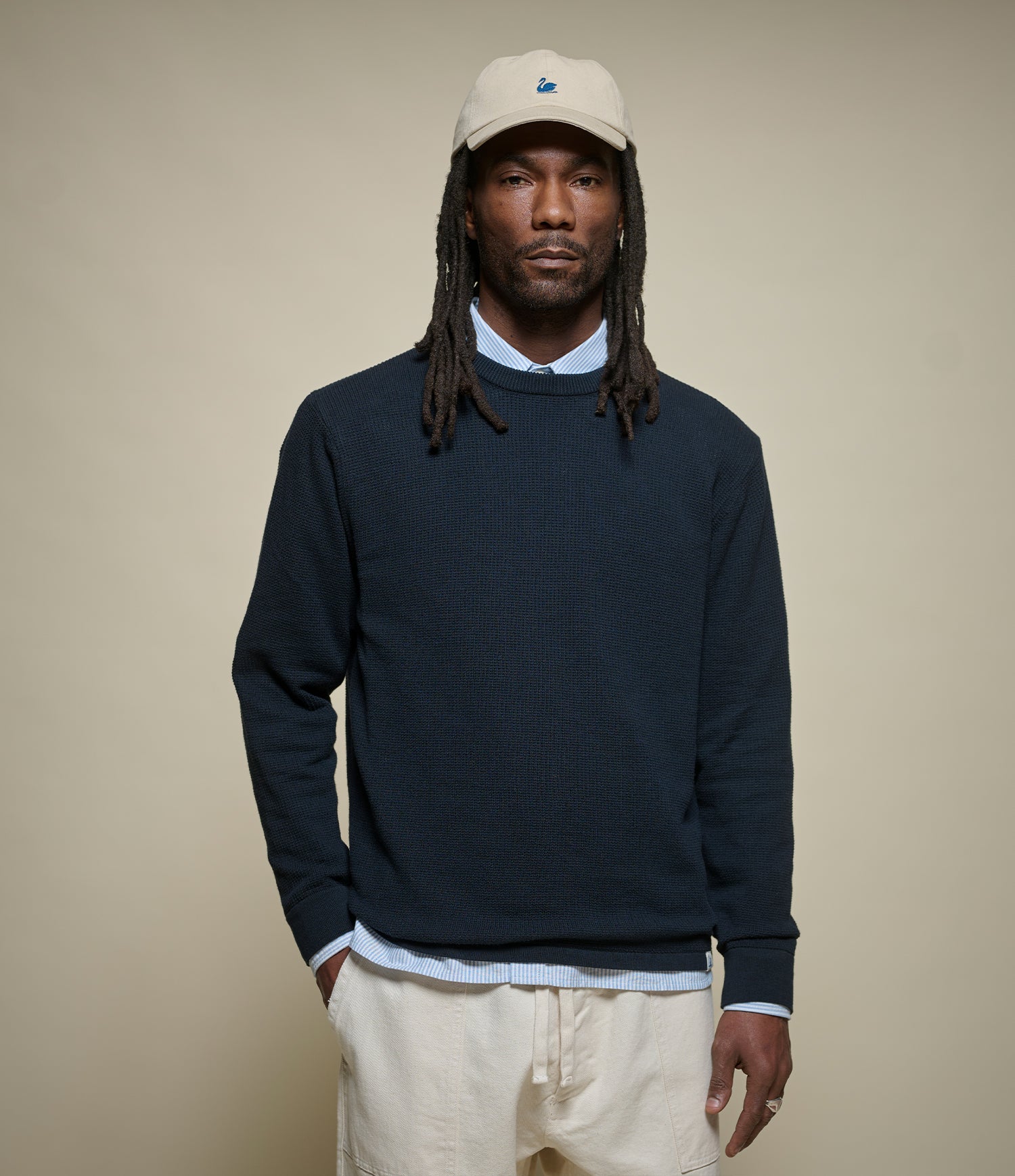 Man wearing a navy lightweight sweater, beige cap, and light-colored pants against a beige background