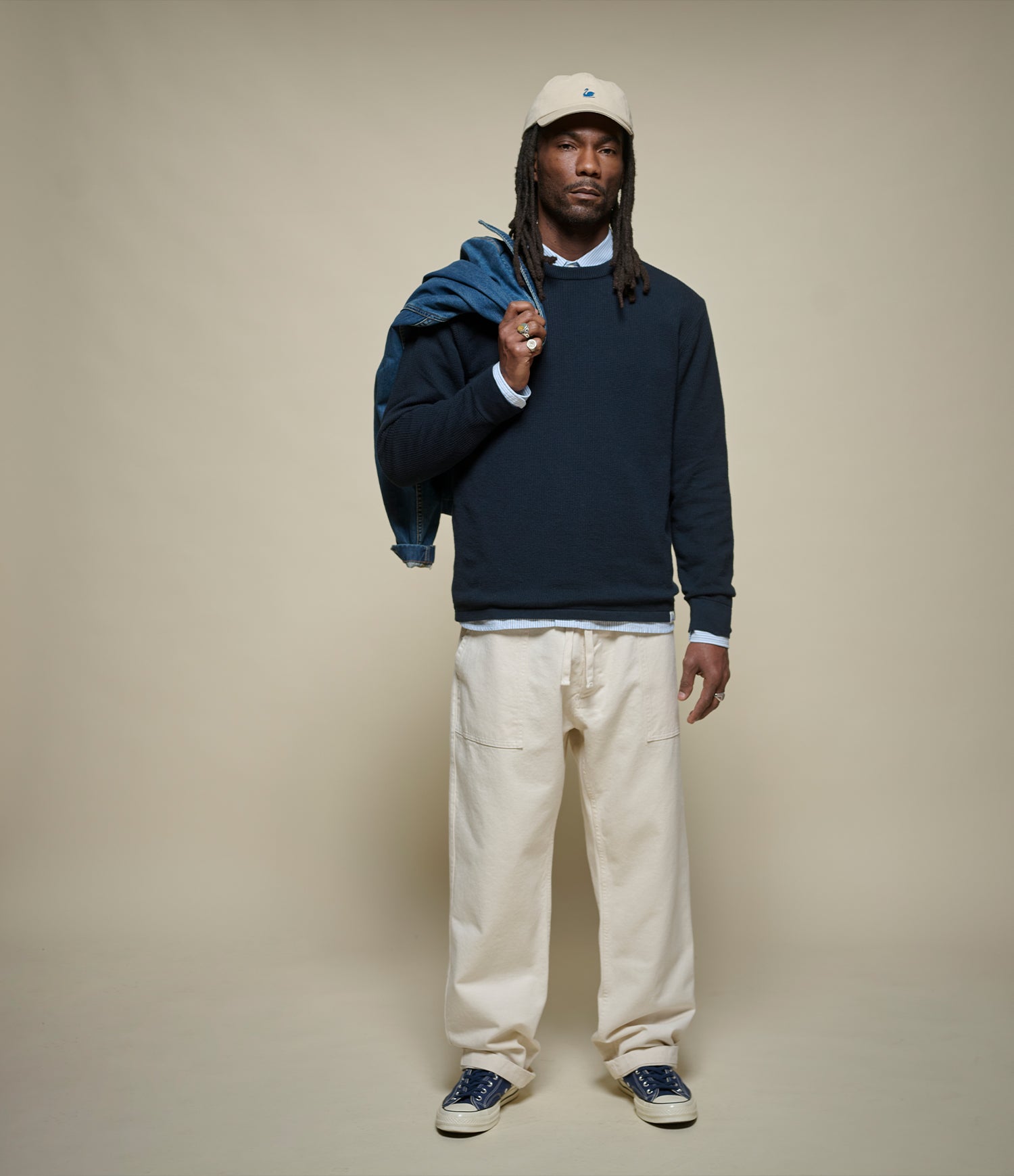 Man wearing a navy sweater, beige pants, and a cap holding a jacket against a beige background