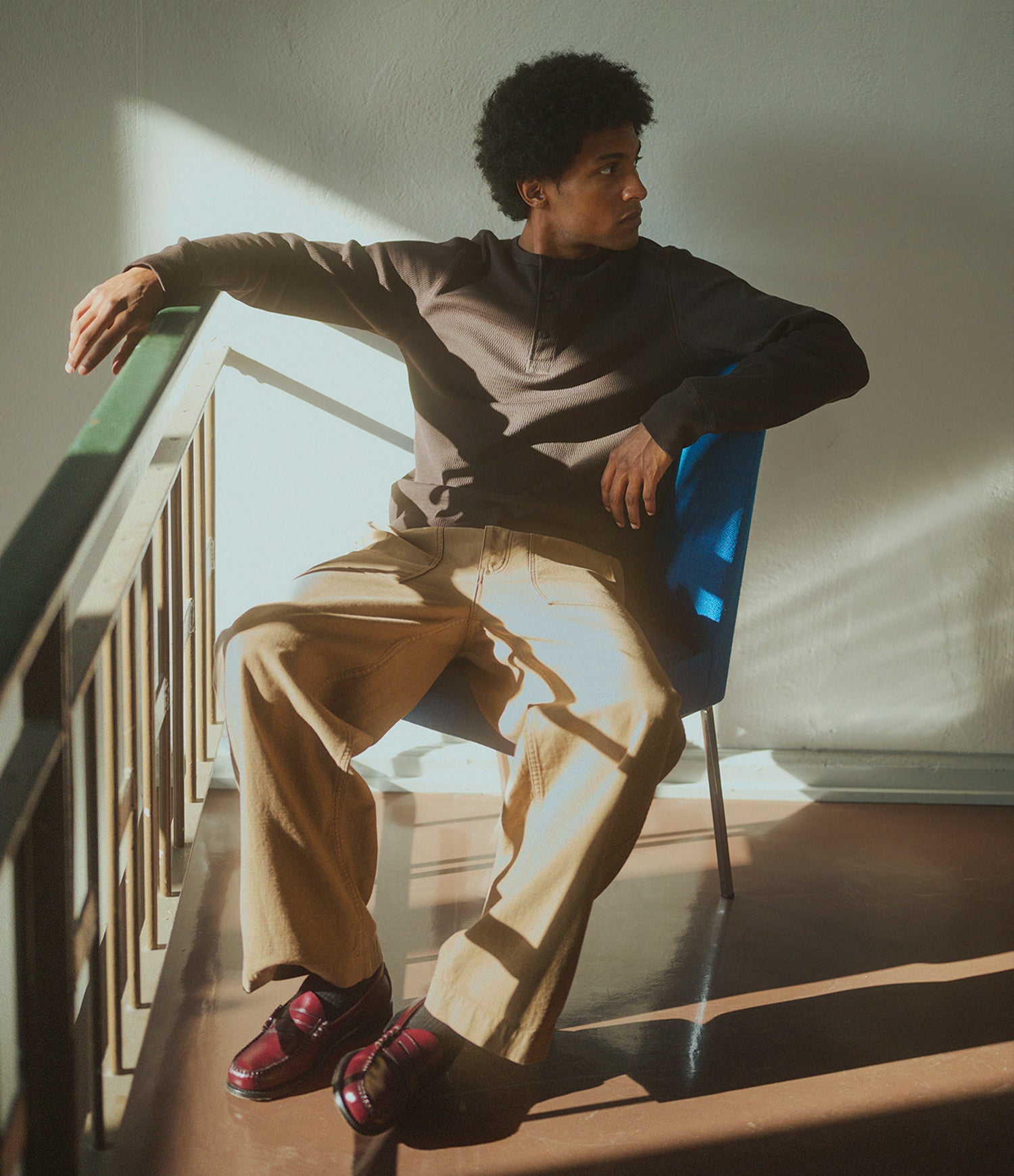 A man sits on a blue chair on an indoor staircase, wearing a dark brown long-sleeve thermal henley, beige wide-leg trousers, and burgundy loafers. Strong sunlight casts dramatic shadows on the wall and floor.