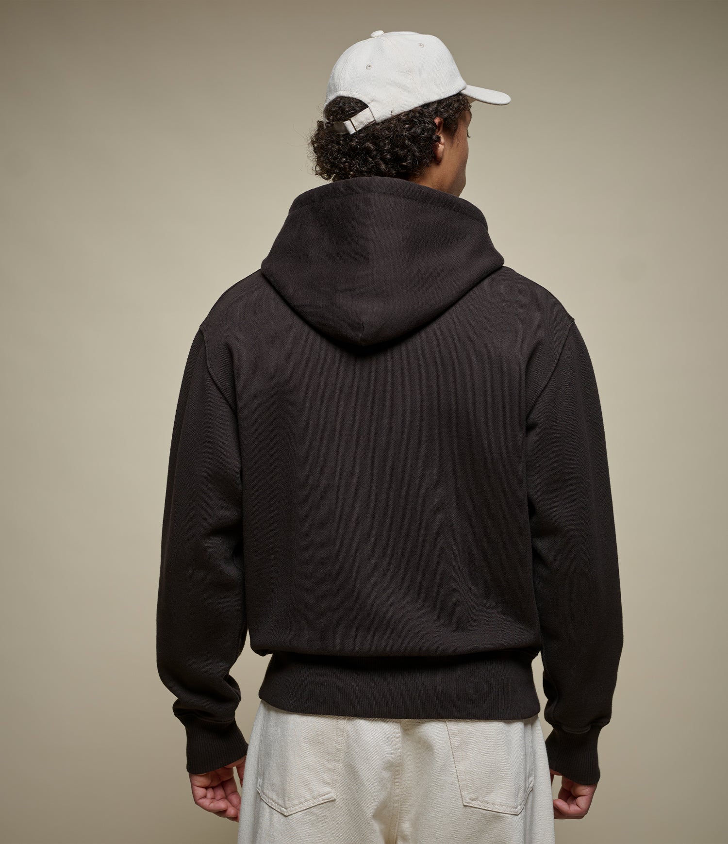 A person shown from the back wearing a dark brown ribbed hoodie, a white baseball cap, and off-white pants against a plain beige studio background.