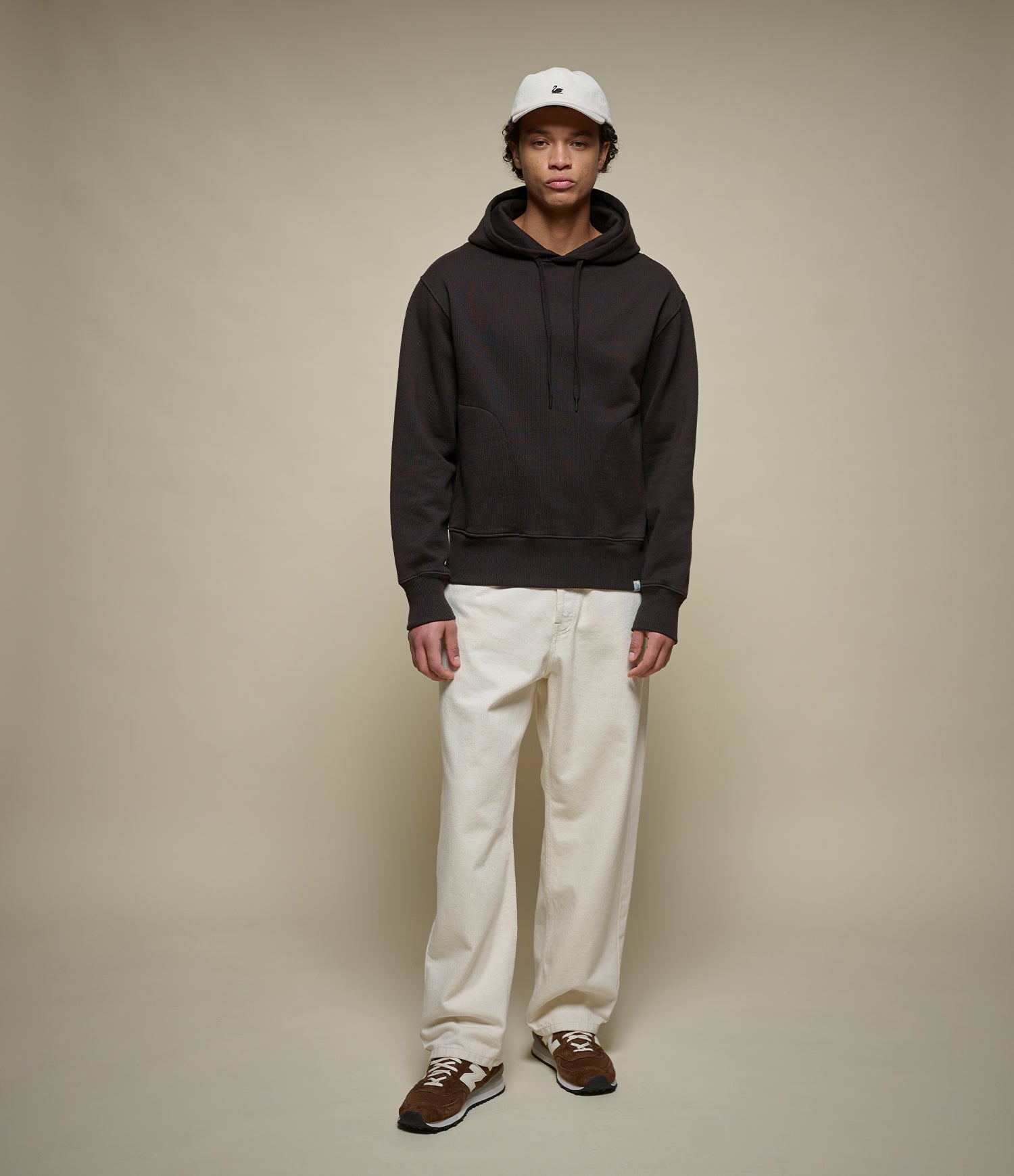 A model stands against a plain beige studio background wearing a dark charcoal hoodie, off-white pants, brown shoes, and a white baseball cap.