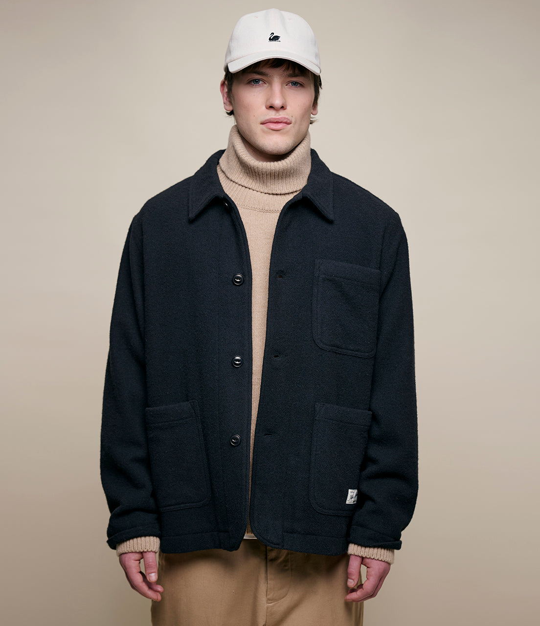 Person wearing a black burel wool jacket, beige turtleneck, and light-colored pants against a beige background