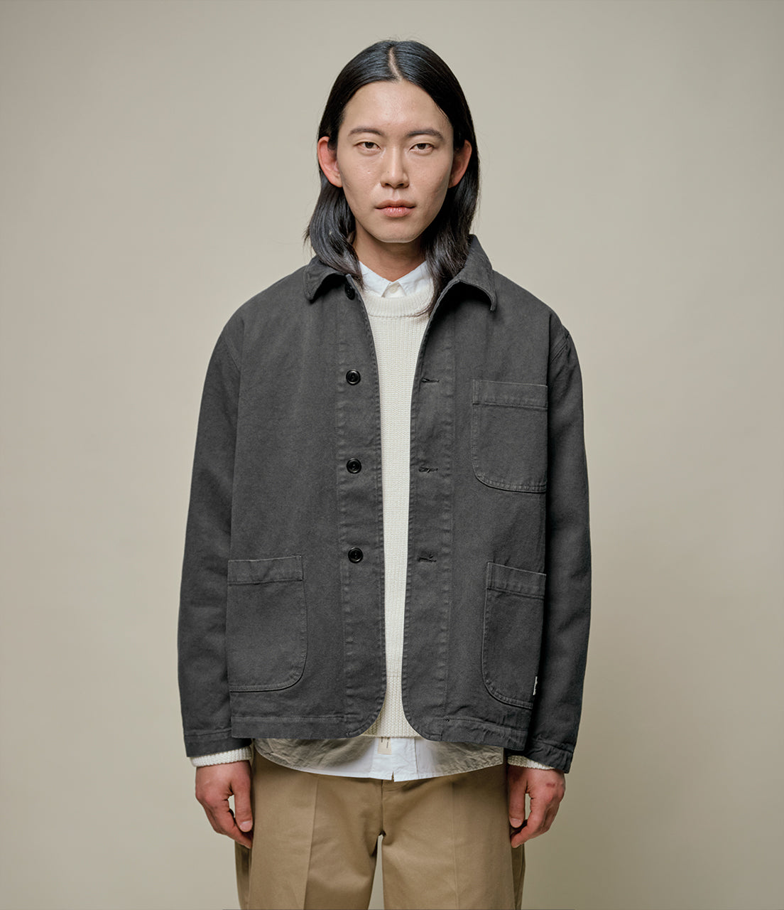 Man wearing a dark gray jacket over a white shirt with beige pants on a plain background
