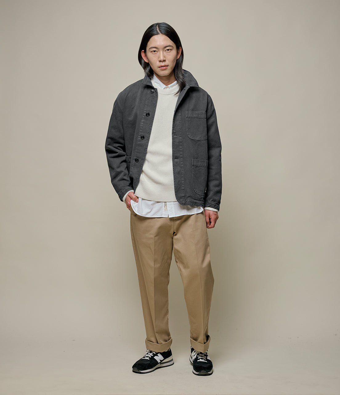 Man wearing a gray jacket, white shirt, beige pants, and black shoes on a plain background