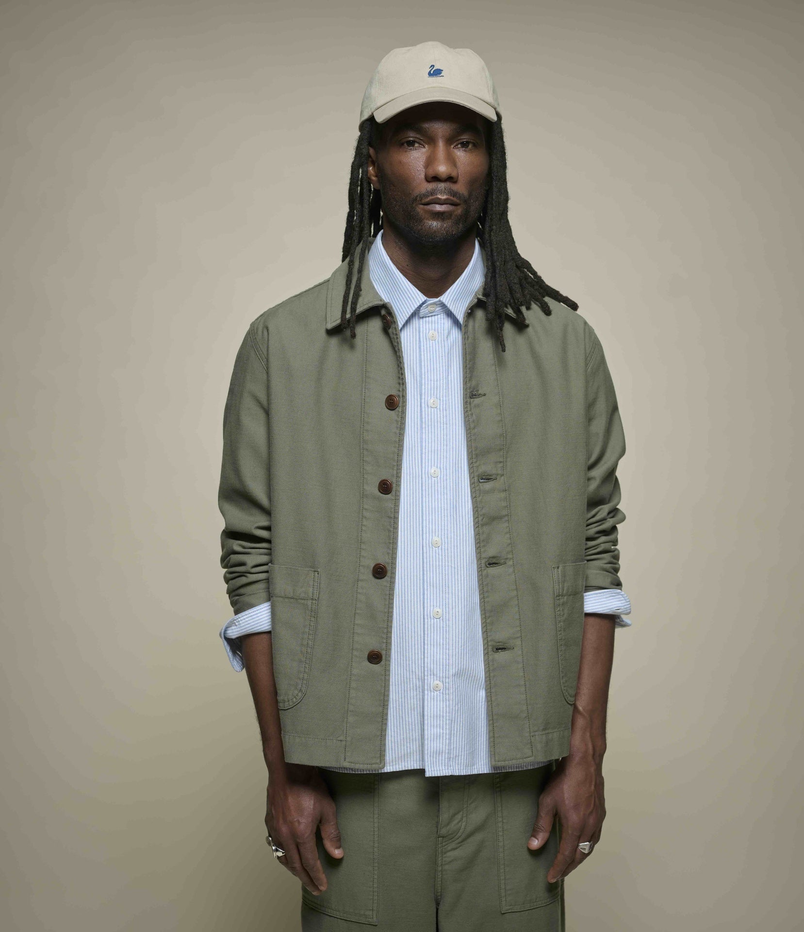 A man wearing a light beige baseball cap, a light blue striped button-down shirt, an olive green utility jacket, and matching olive green pants, standing against a solid beige background.