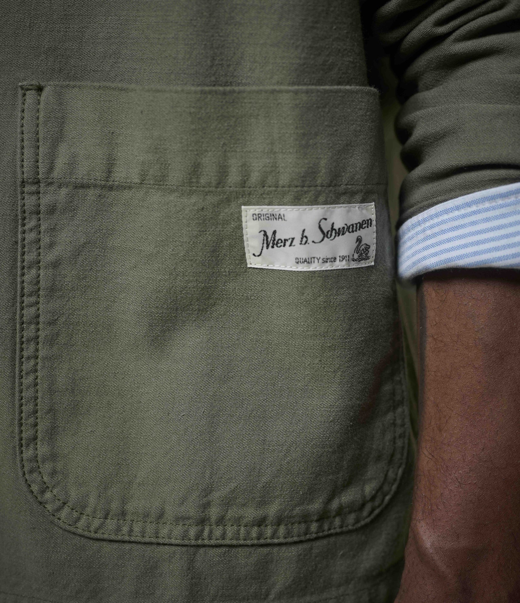 Close-up of an olive green fabric pocket with a white sewn-in label reading 'Merz b. Schwanen' and a striped shirt cuff visible on the right.