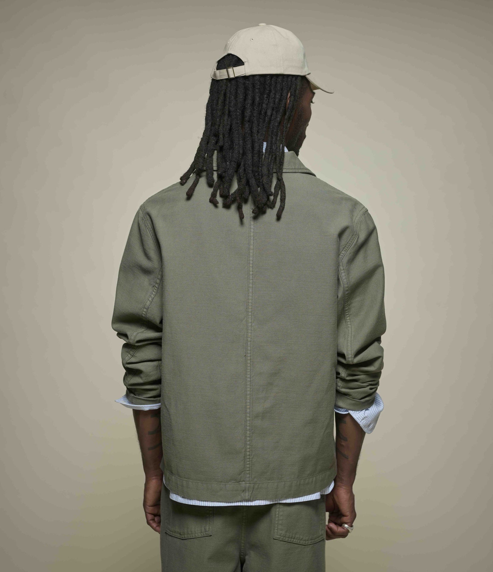 A man viewed from behind wearing an olive green canvas utility jacket, matching green pants, a white cap, and a white long-sleeve shirt underneath, set against a plain beige studio background.