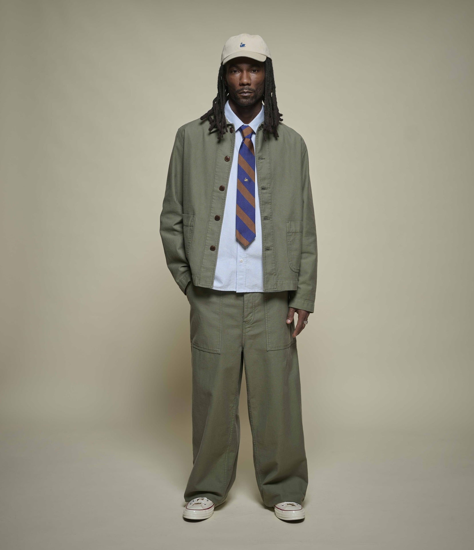 A model stands against a plain beige studio background wearing a matching olive green two-piece suit set consisting of a button-up jacket and wide-leg trousers, styled with a white shirt, a blue and orange striped tie, and a white cap.