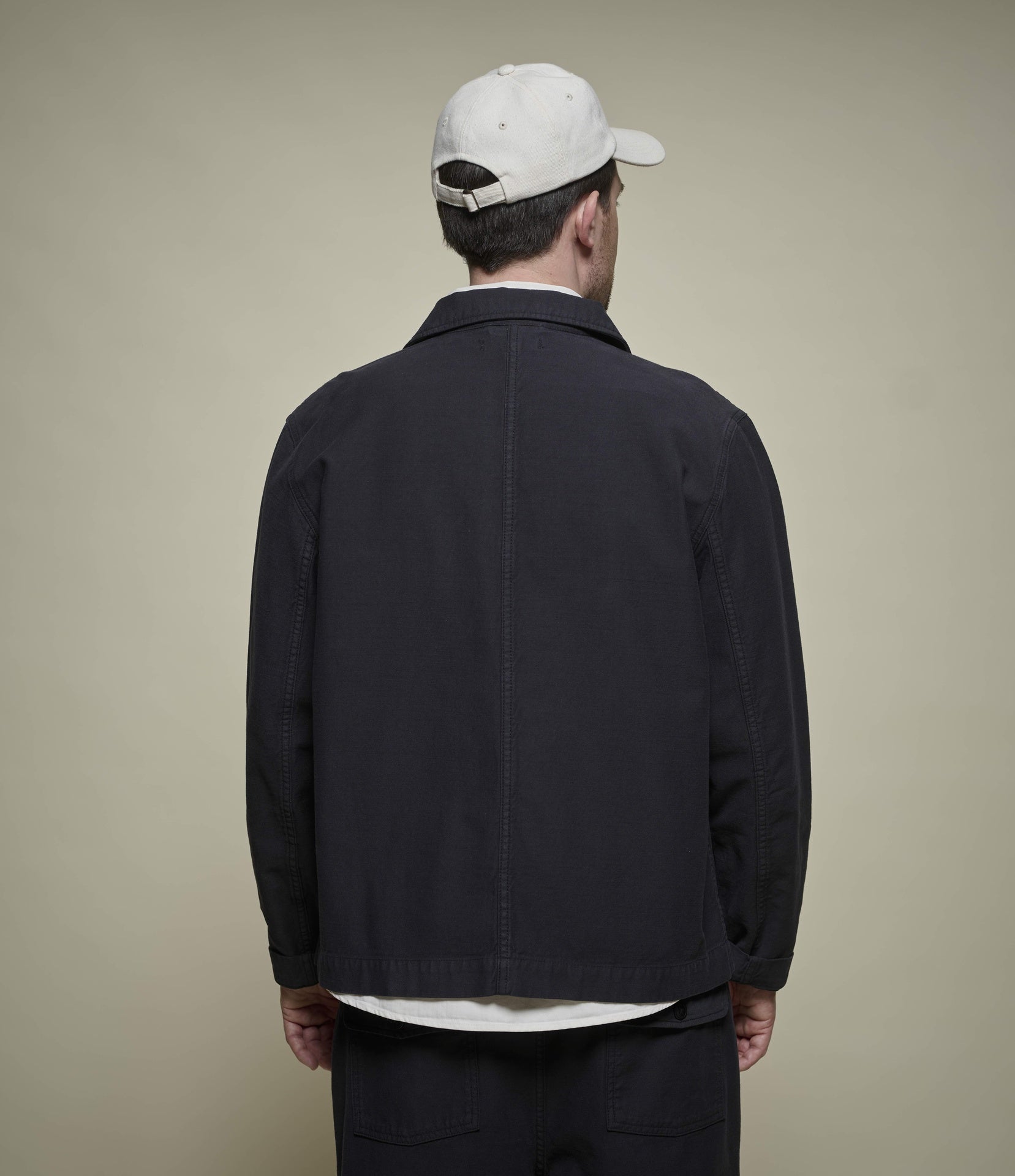 A man viewed from behind wearing a dark charcoal utility jacket, a white t-shirt, and a white baseball cap against a solid beige background.