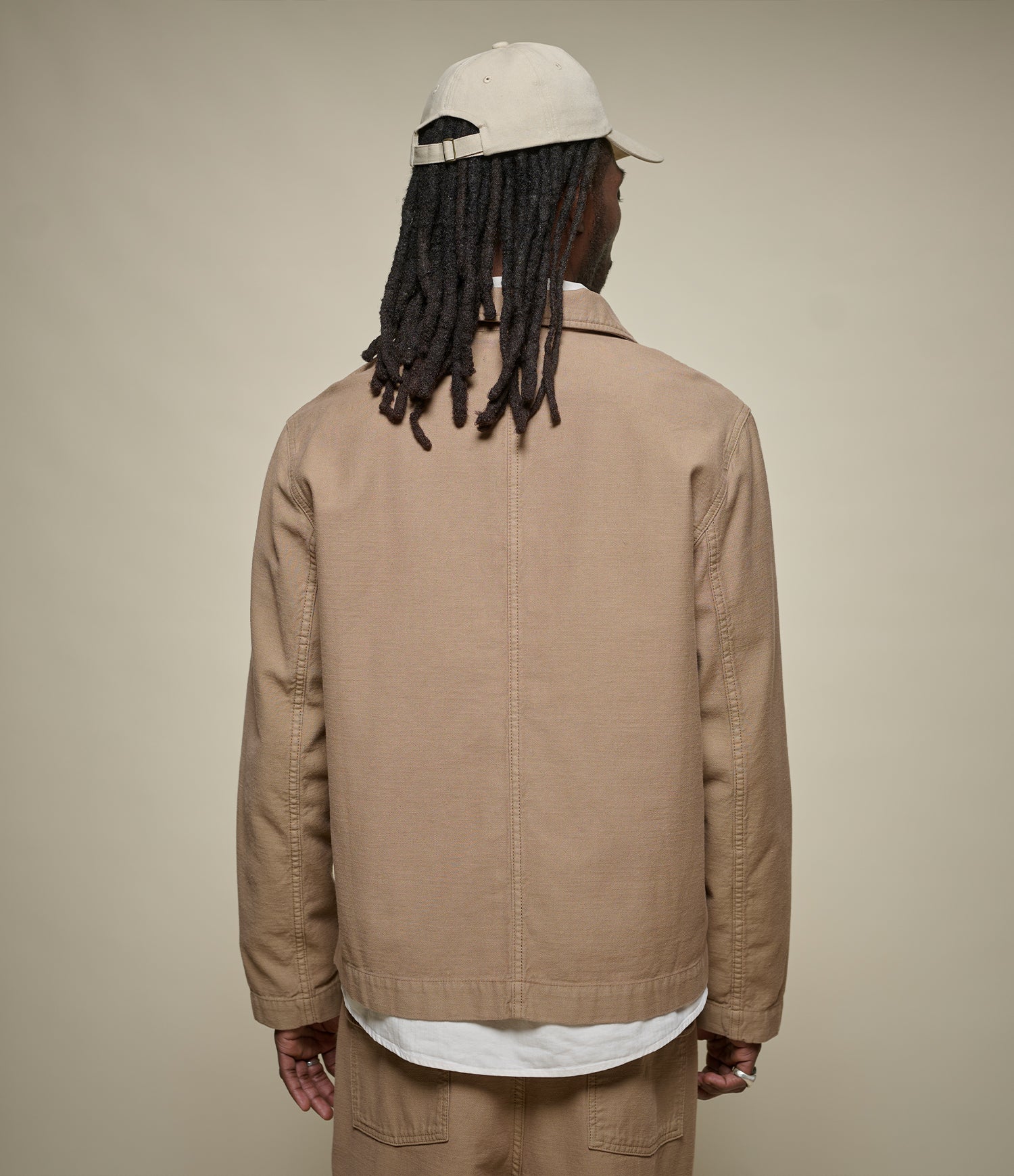 A man viewed from behind wearing a beige utility jacket, matching beige pants, a white t-shirt, and a white baseball cap against a plain beige studio background.