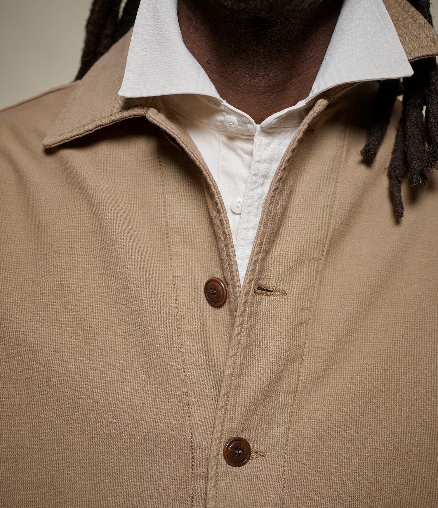 Close-up of a person wearing a tan sateen canvas jacket over a white collared shirt. The tan garment has a collar, front button placket, and visible seam detailing, with dark brown buttons.