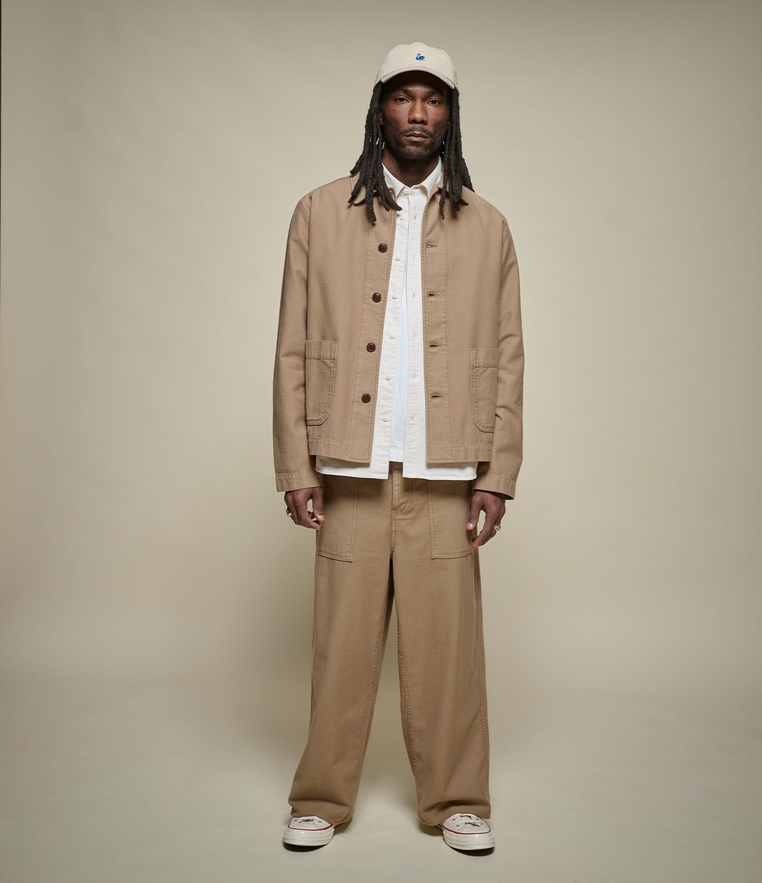 A model stands against a plain beige studio background wearing a beige utility jacket and matching wide-leg trousers over a white shirt, with a white cap and white sneakers.