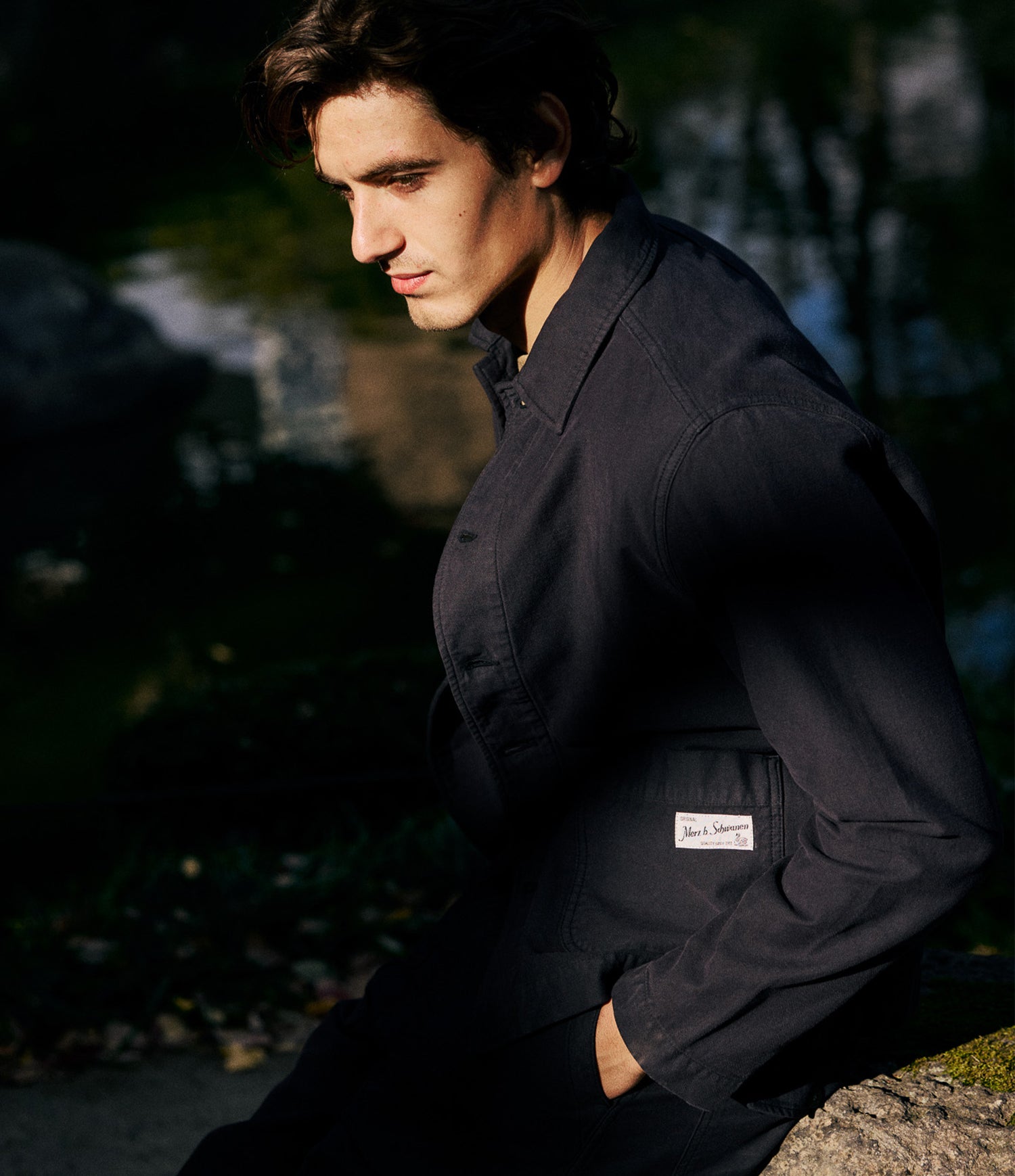 A man wearing a dark charcoal gray work jacket with a visible Merz b. Schwanen label on the pocket, posed outdoors in dramatic, low-key lighting against a dark, blurred natural background.