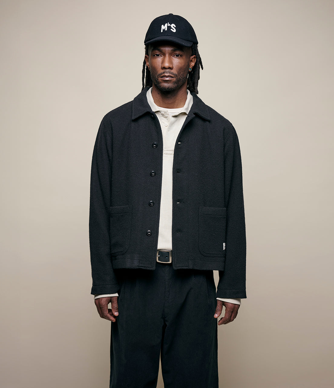 Man wearing a black burel wool jacket, white shirt, and cap on a beige background