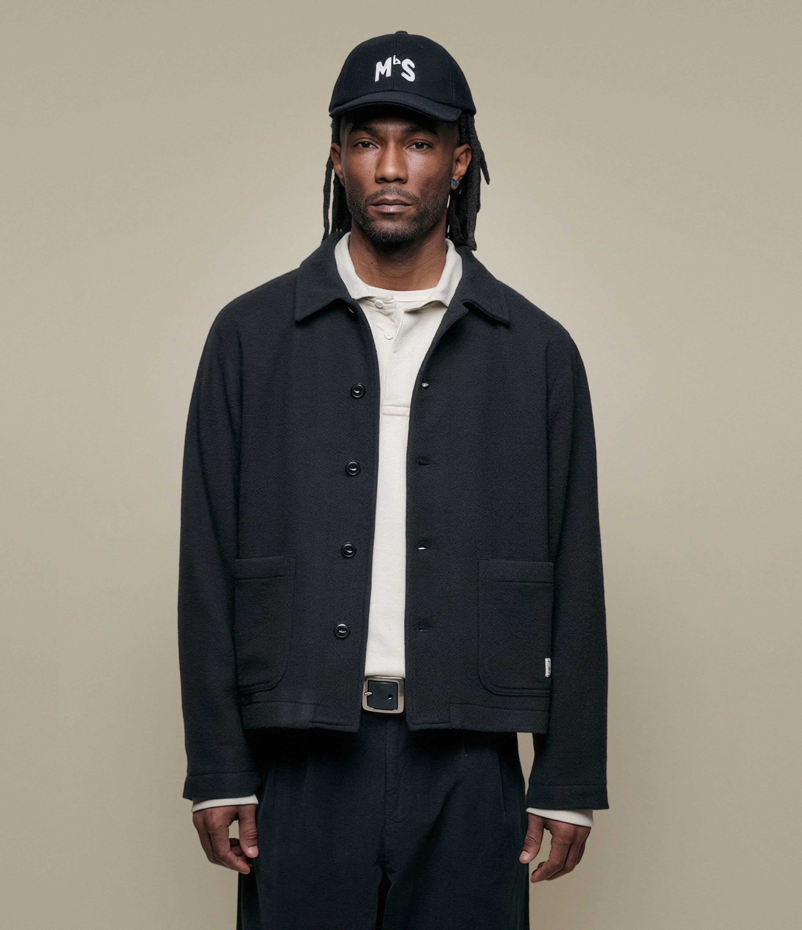 Man wearing a black burel wool jacket, white shirt, and cap on a beige background