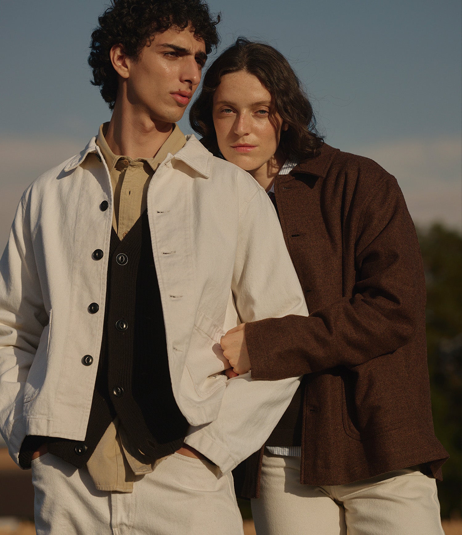 Two models standing outdoors wearing layered vintage-style clothing. The model on the left wears an off-white utility jacket over a black vest and beige shirt, while the model on the right wears a brown textured jacket over a dark top.
