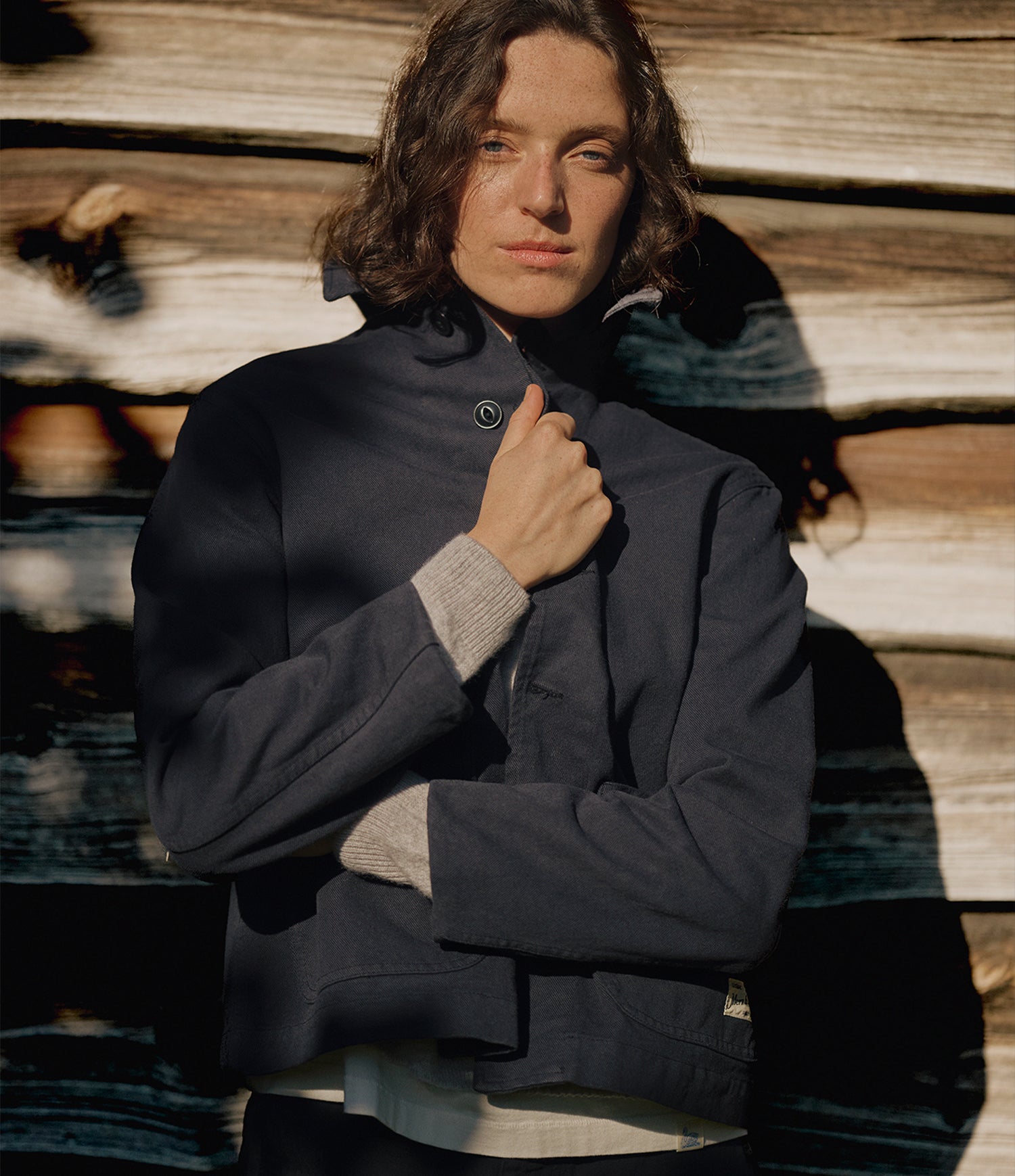 A model with shoulder-length brown hair stands against a rustic wooden wall, wearing a dark navy grey cropped jacket with a high collar and a light gray knit sweater layered underneath.