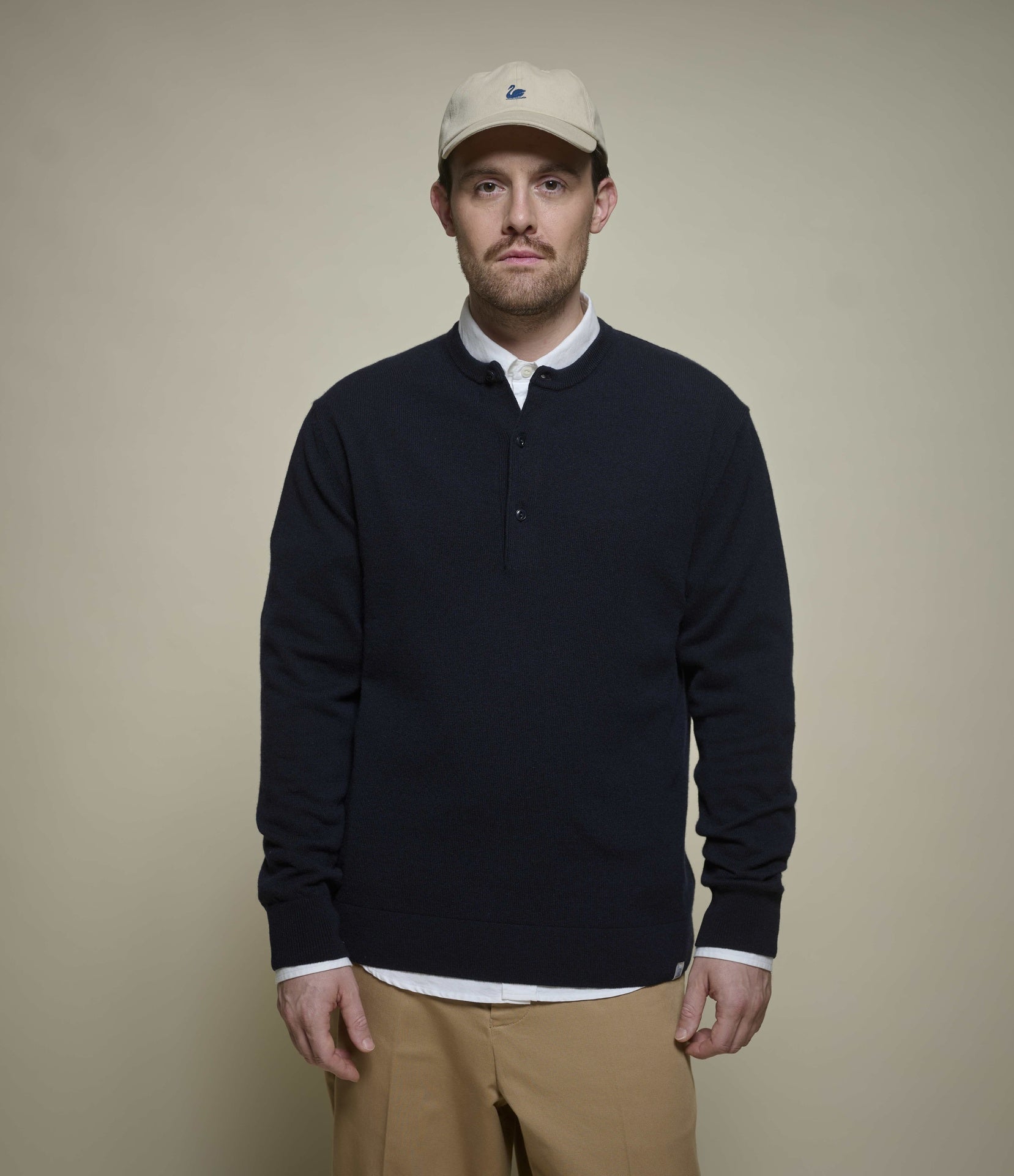 A man wearing a dark navy long-sleeve henley shirt over a white collared undershirt, khaki pants, and a beige baseball cap, standing against a solid beige background.